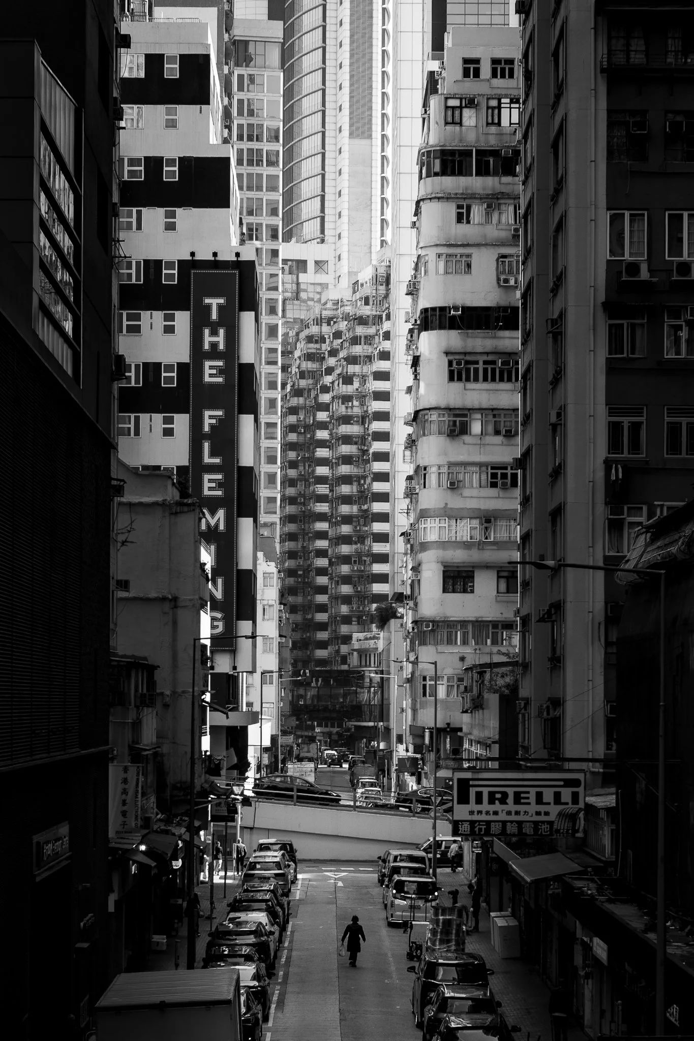 A narrow city street filled with parked cars, with high-rise buildings on both sides and a woman walking in the center. Signage in English and Chinese is visible on the buildings, including a large sign with the word "THE FILMING" and another adverti
