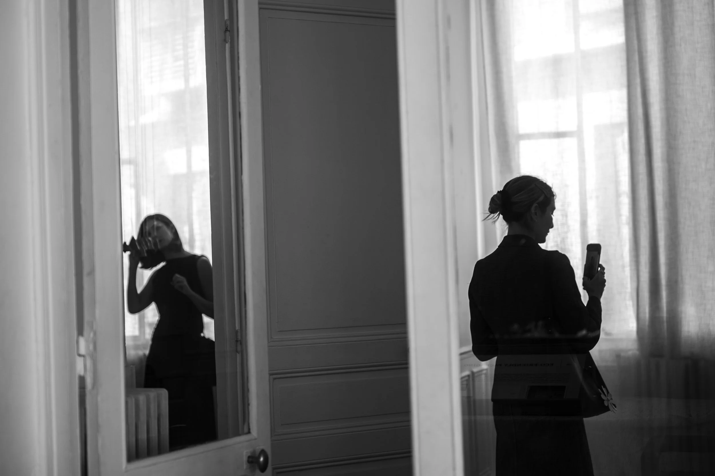 A woman with her hair tied up looking at her phone, seen through a reflection in a glass pane, with sheer curtains and window blinds in the background.