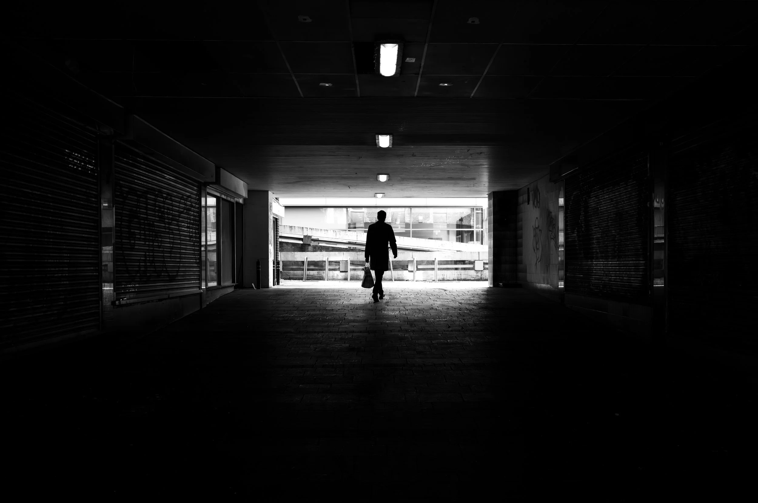 Black and white photo of a man walking under a tunnel, carrying shopping bags, with bright light and building exterior visible at the end of the tunnel.