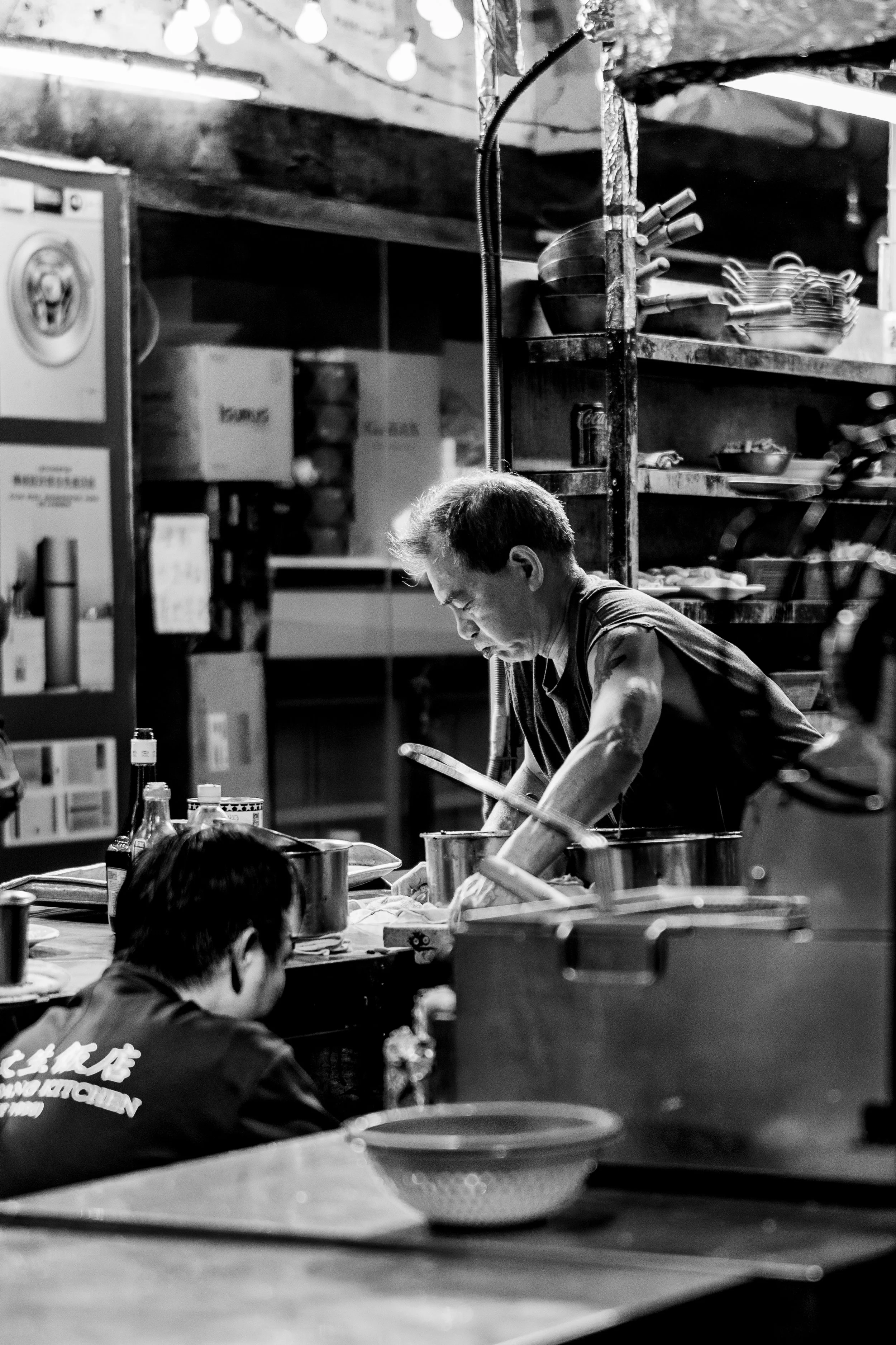 A black and white photo of a person cooking or preparing food in a kitchen or restaurant setting. Several people are visible, with one man focused on his task, surrounded by bowls, utensils, and kitchen supplies.