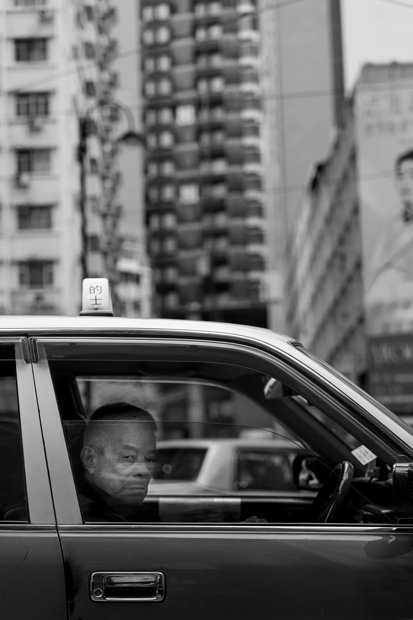A man with gray hair and a face mask looking out of the passenger side window of a pickup truck in front of tall apartment buildings.