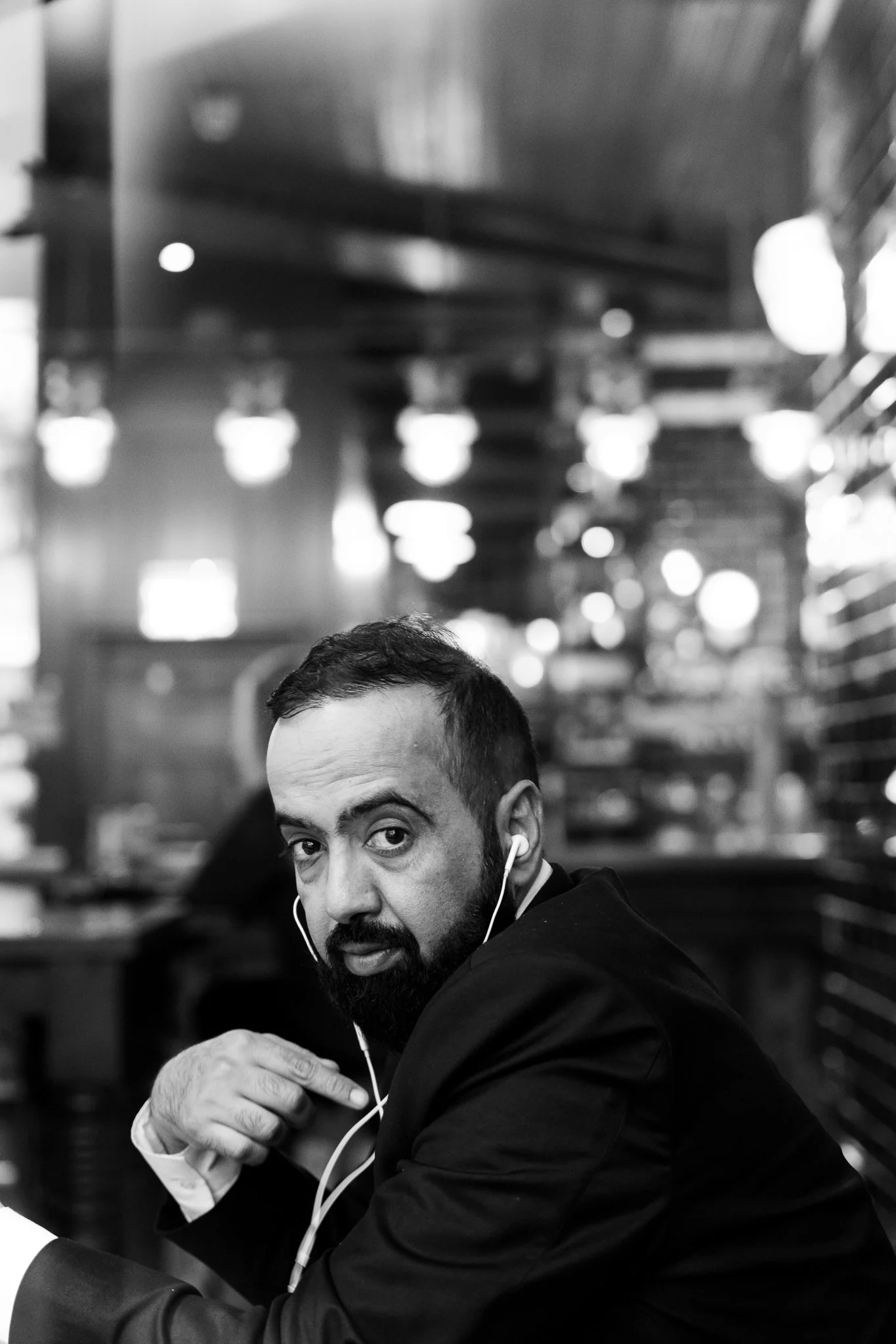 A man with short hair and a beard wearing a suit and airpods, looking over his shoulder in a modern, well-lit restaurant or cafe.