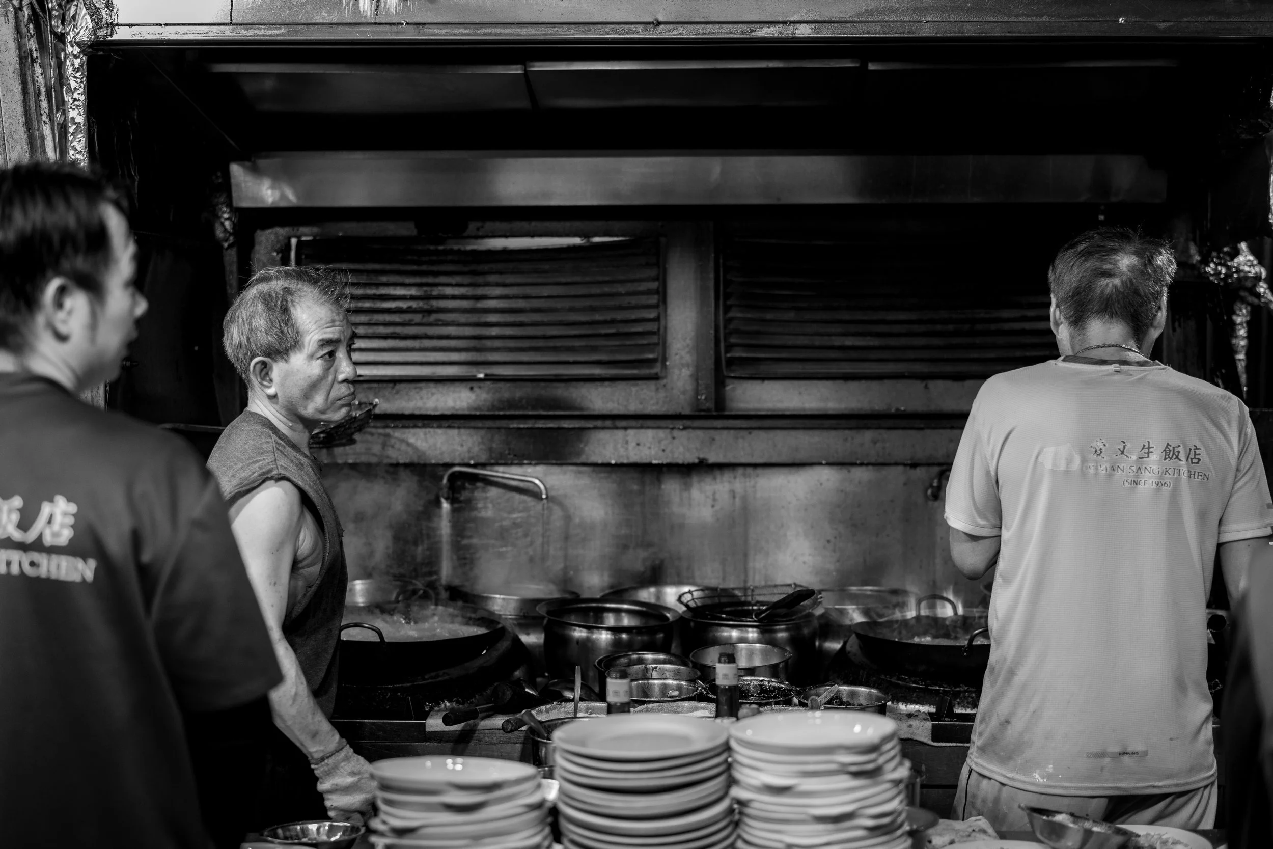 Four men working in a busy restaurant kitchen, preparing food behind the counter. Five stacks of white bowls are in the foreground.