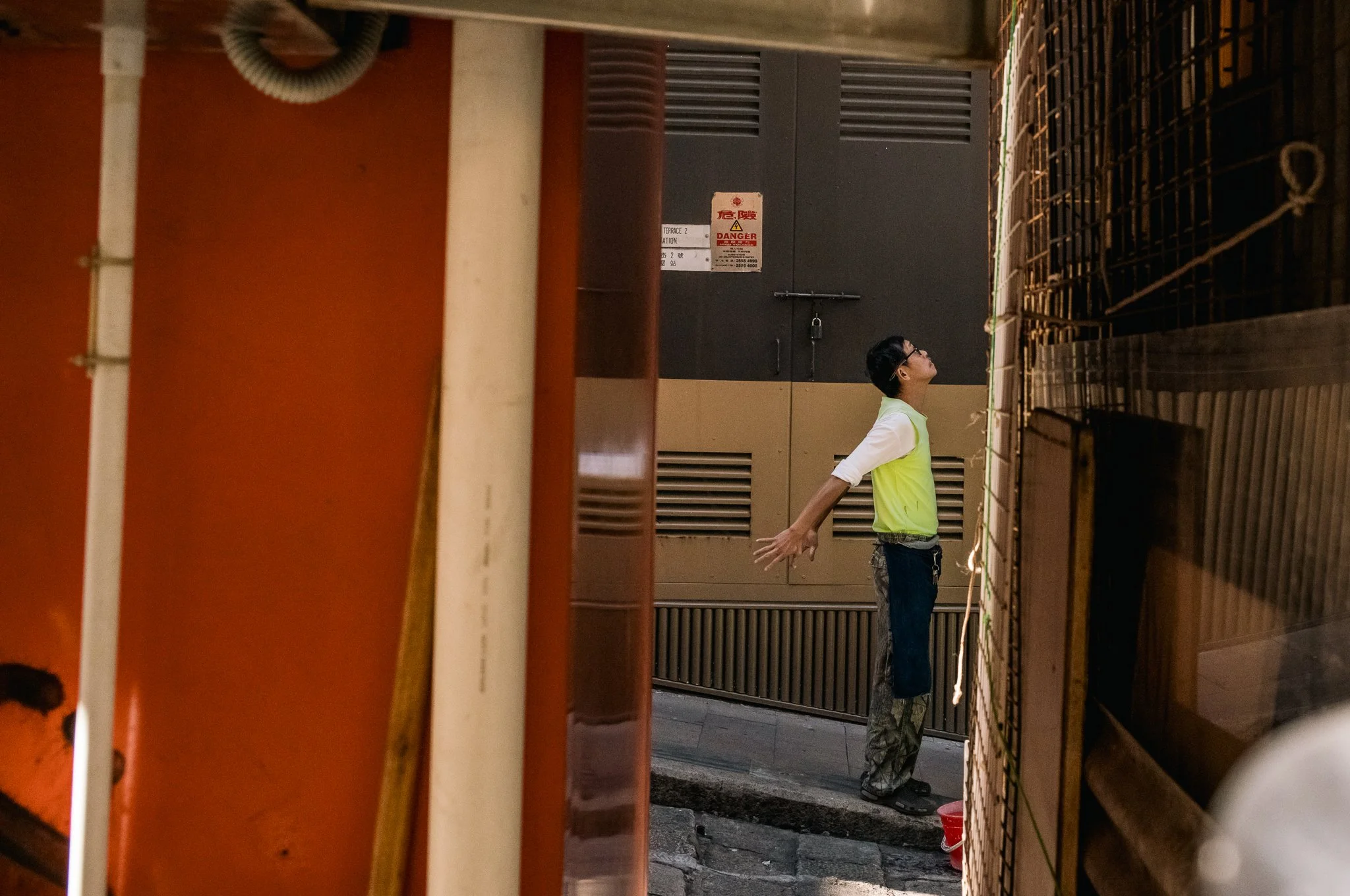 A man in a yellow vest with his arms outstretched stands in an alleyway between buildings, seen through a narrow gap.