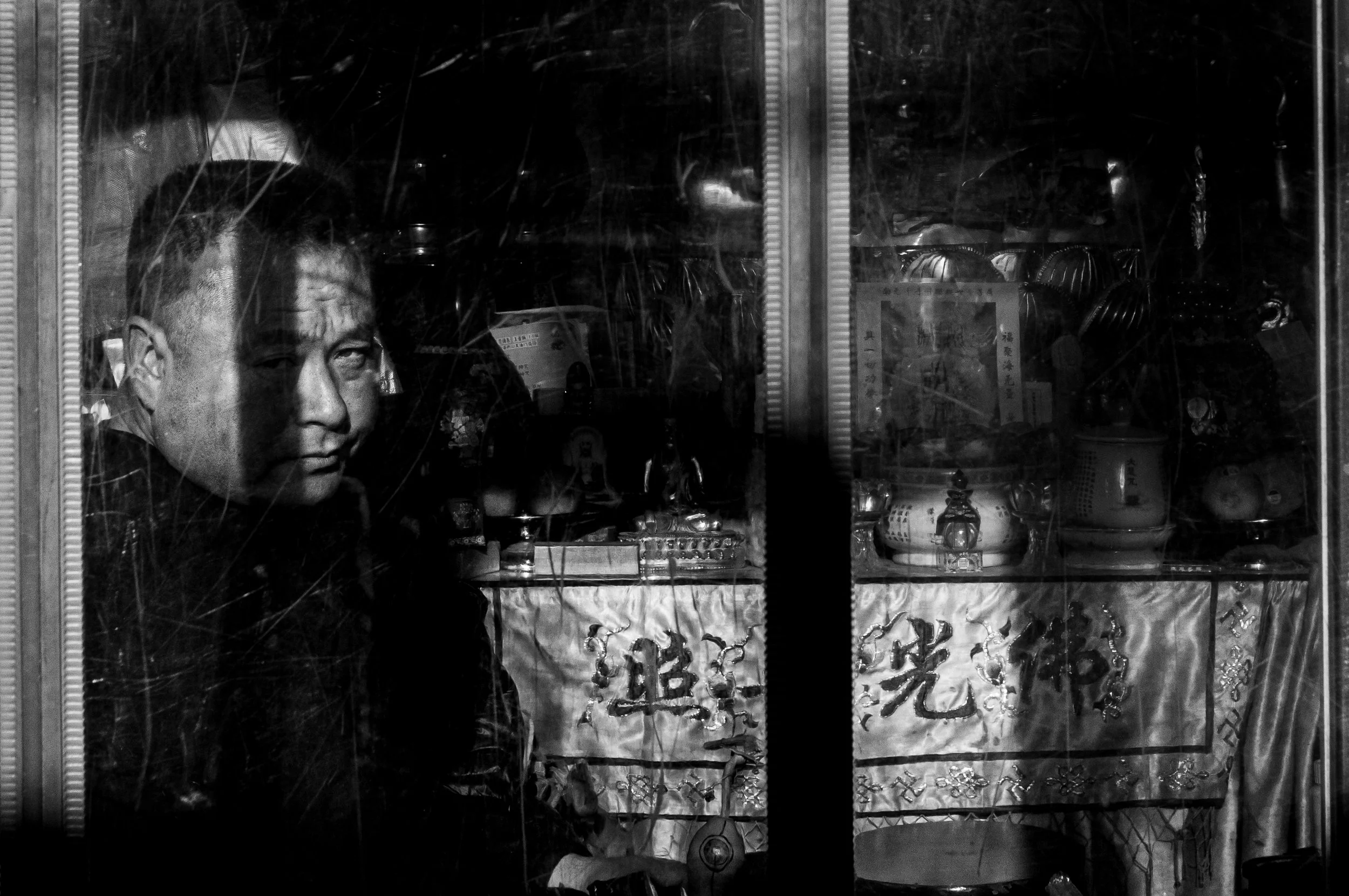 A person with short hair and a serious expression looking through a window with reflections and cracks. Inside the window, there are various objects and decorations on display, including sculptures, ornaments, and Asian script on a sign.