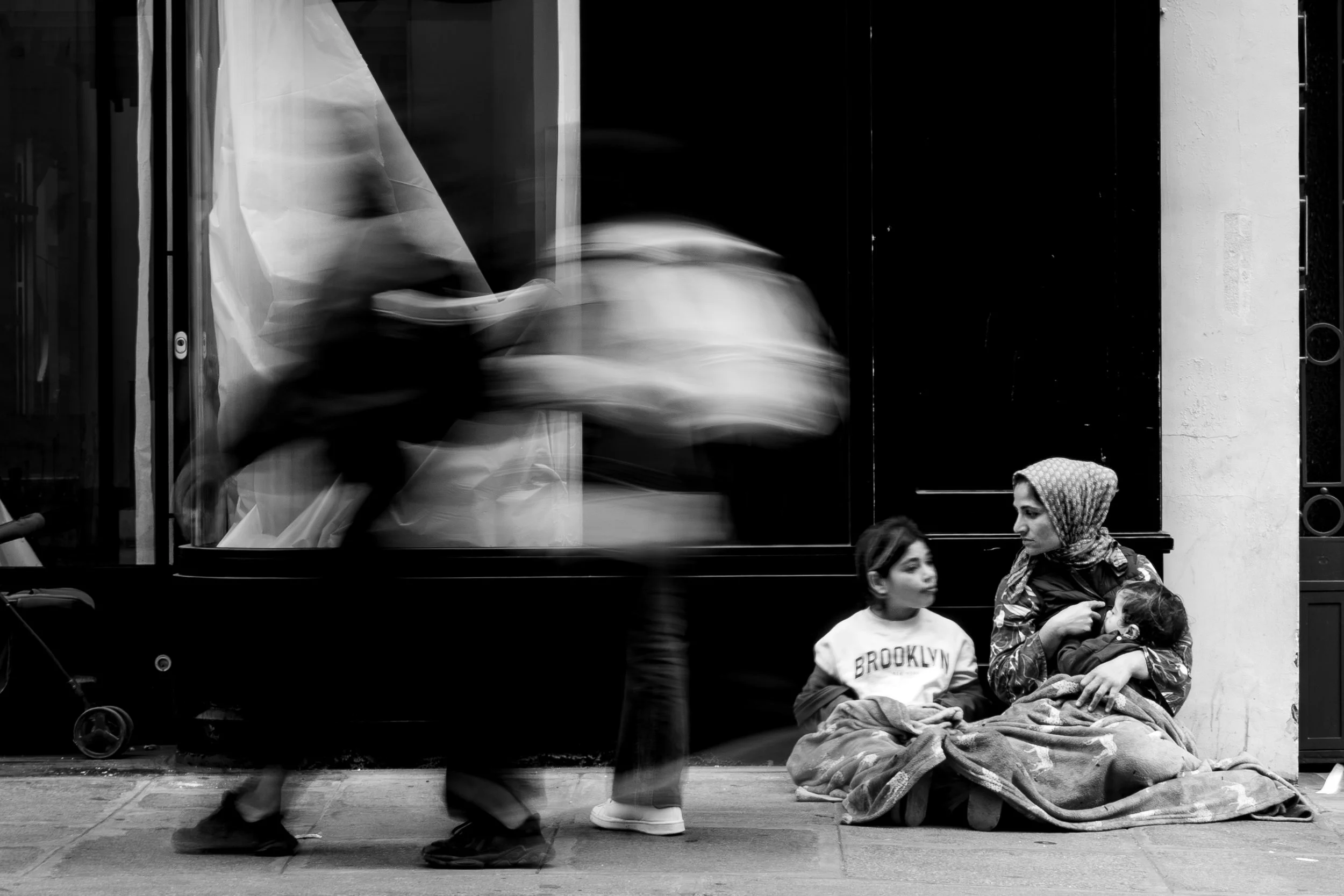 Black and white photo of a city street with blurred people walking past a retailer display window. Two women sit on the sidewalk; one woman is wearing a hijab and holding a child, and a young girl stands in front of them.