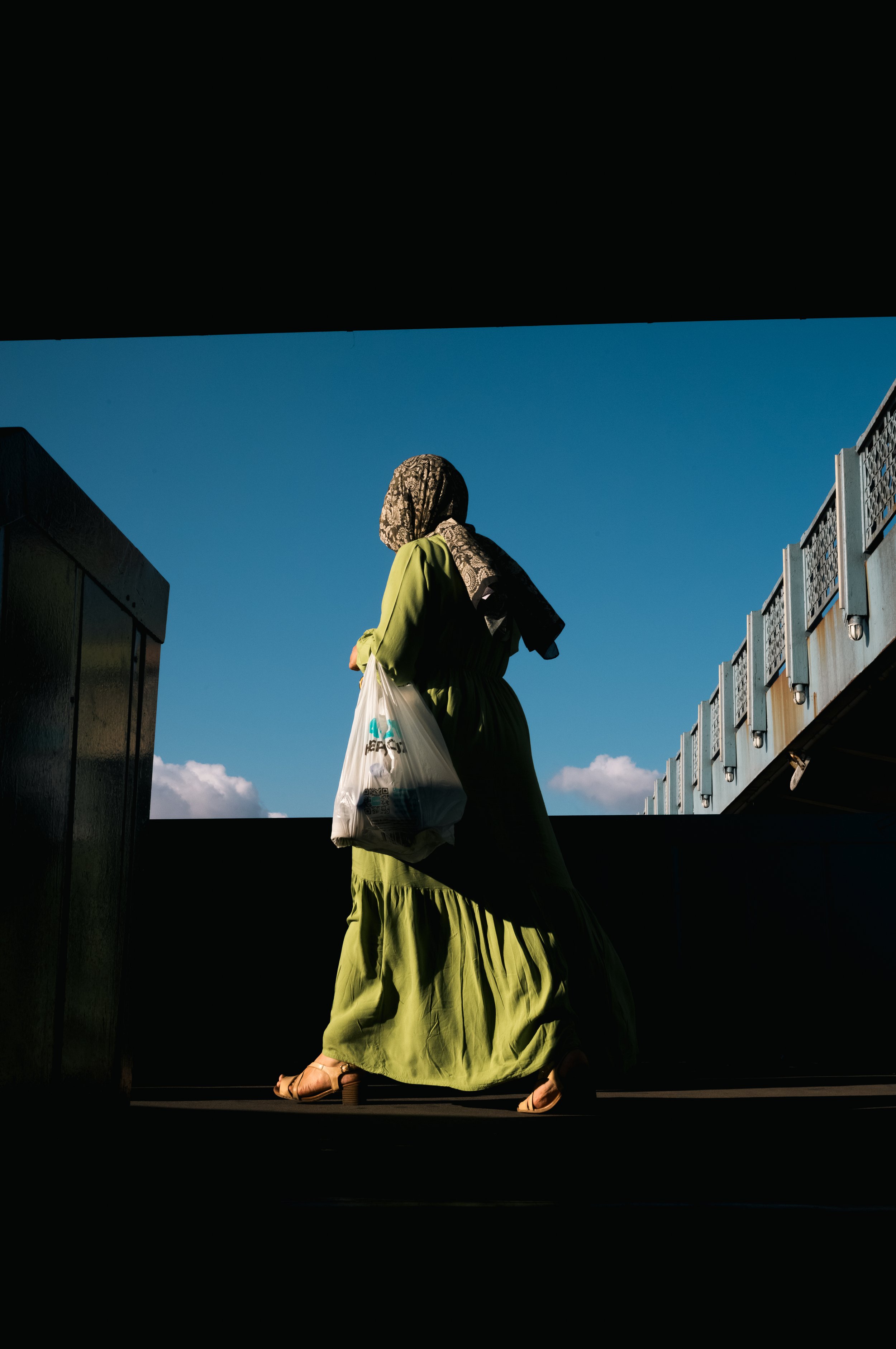 A woman wearing a green dress and a patterned headscarf walks with a plastic bag in hand, seen from a low angle against a bright blue sky, framed by dark shadows and railing.