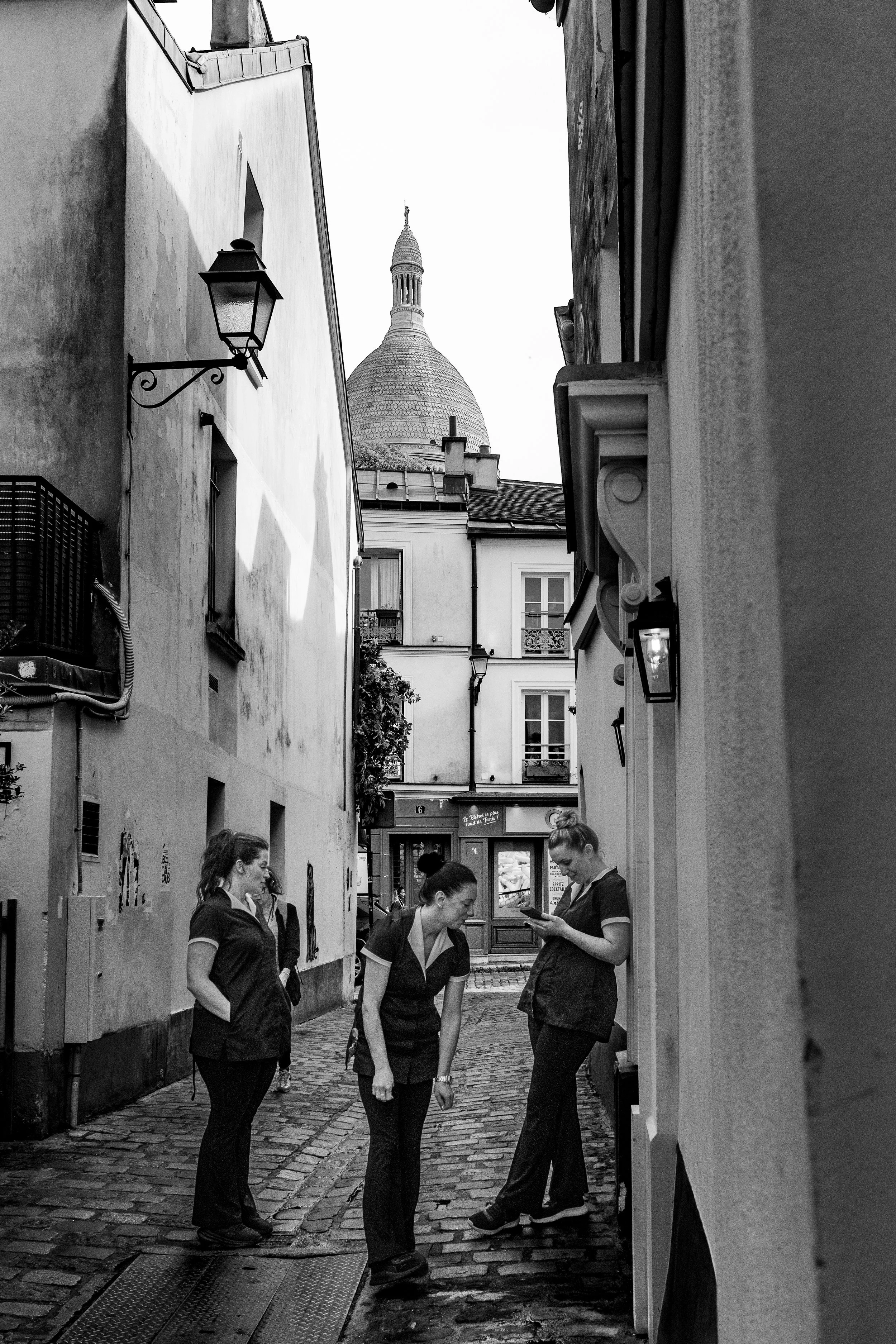 Four women in uniforms standing on a cobblestone street in a European city, with one woman looking at her phone. A dome is visible in the background.