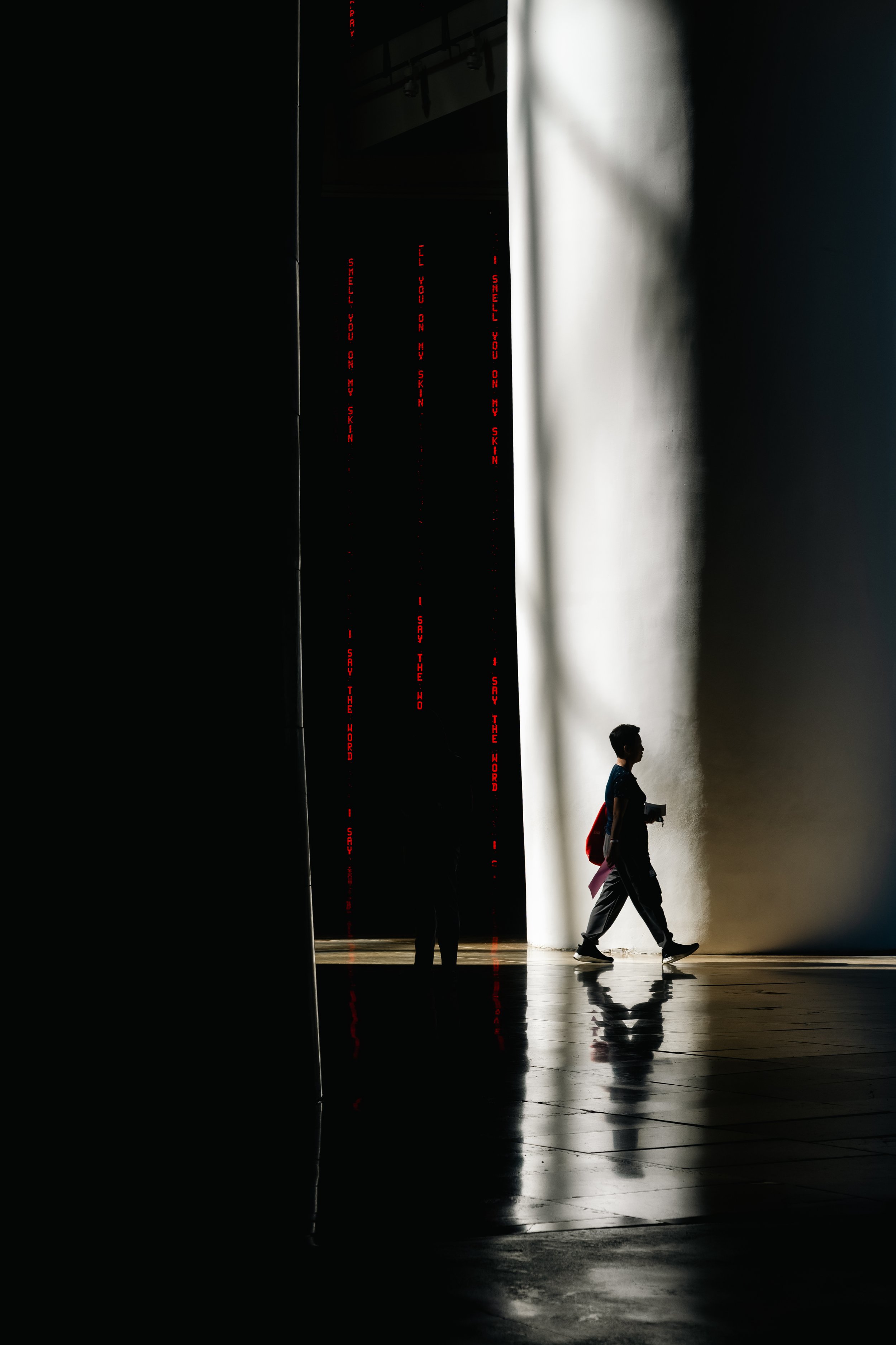A person walking in a corridor with a high contrast light and dark background, with red illuminated text on the dark wall side.