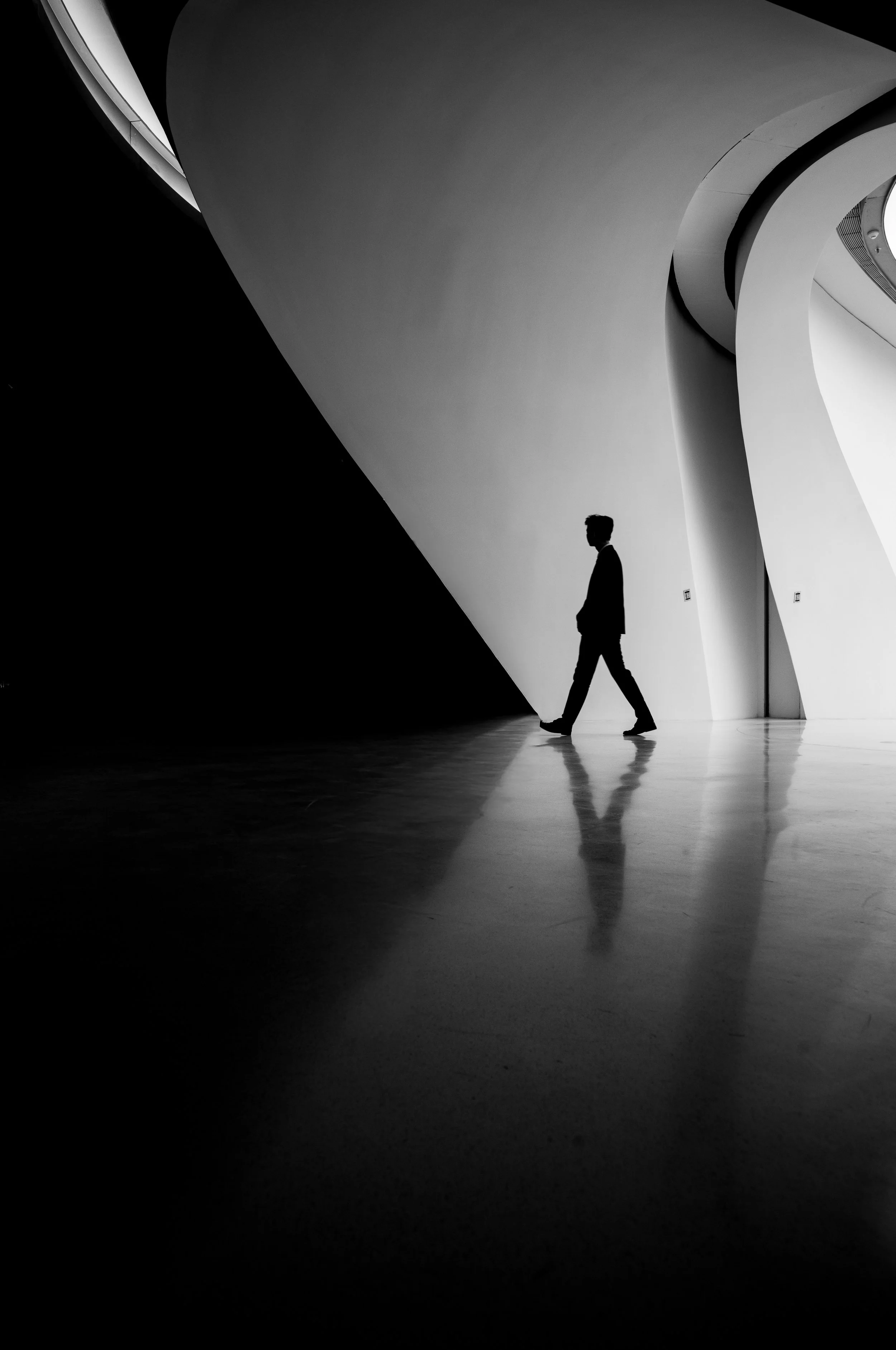 A black and white photo of a man walking in an art gallery with modern architecture. The man is dressed in a suit and is walking on a shiny floor. The gallery has curved walls and distinctive architectural shapes.