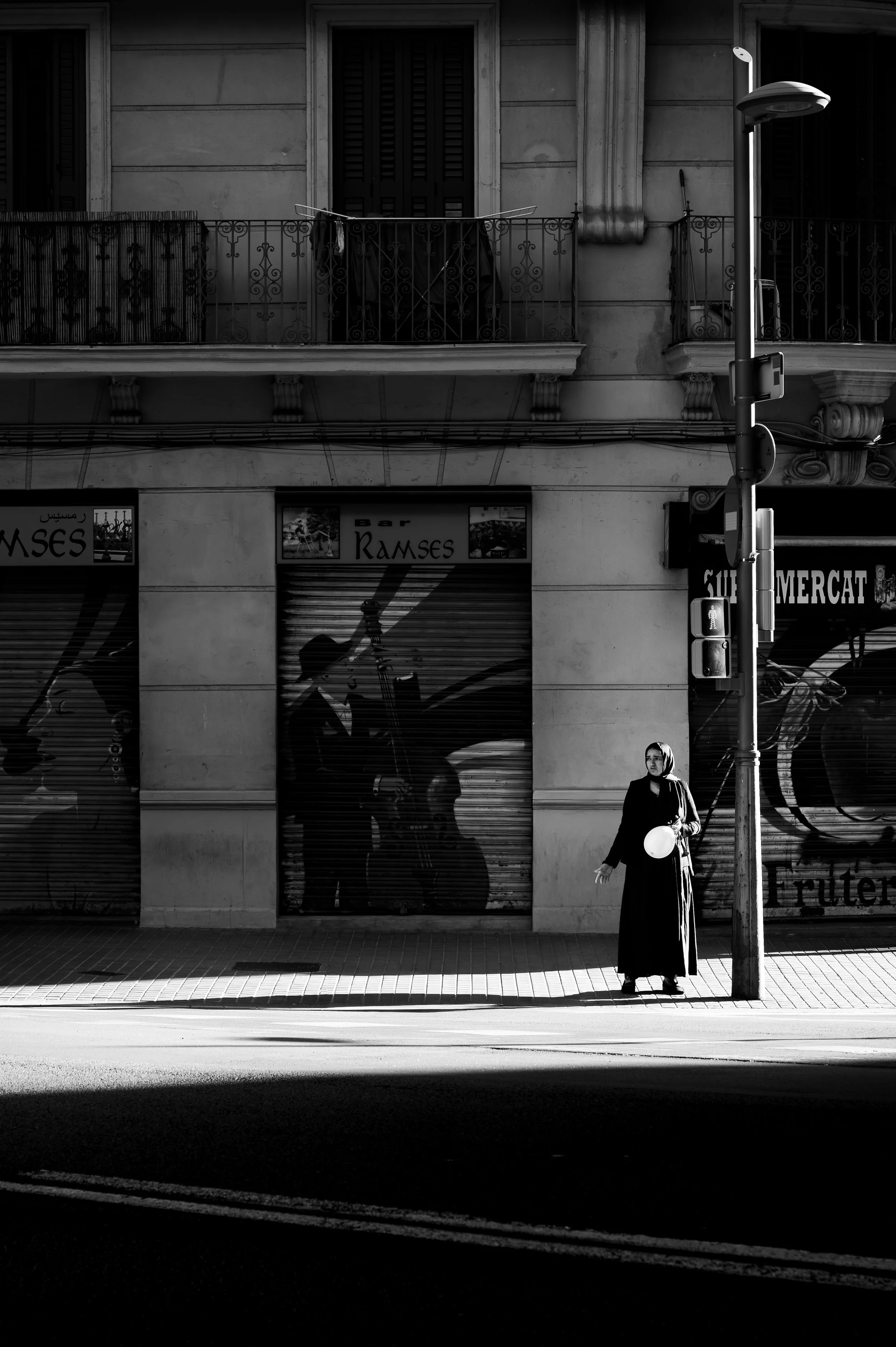 A woman dressed in black holding a white balloon, walking on the sidewalk in front of a building with shutters, balconies, and graffiti-covered roller shutters. A traffic light is visible in the foreground.