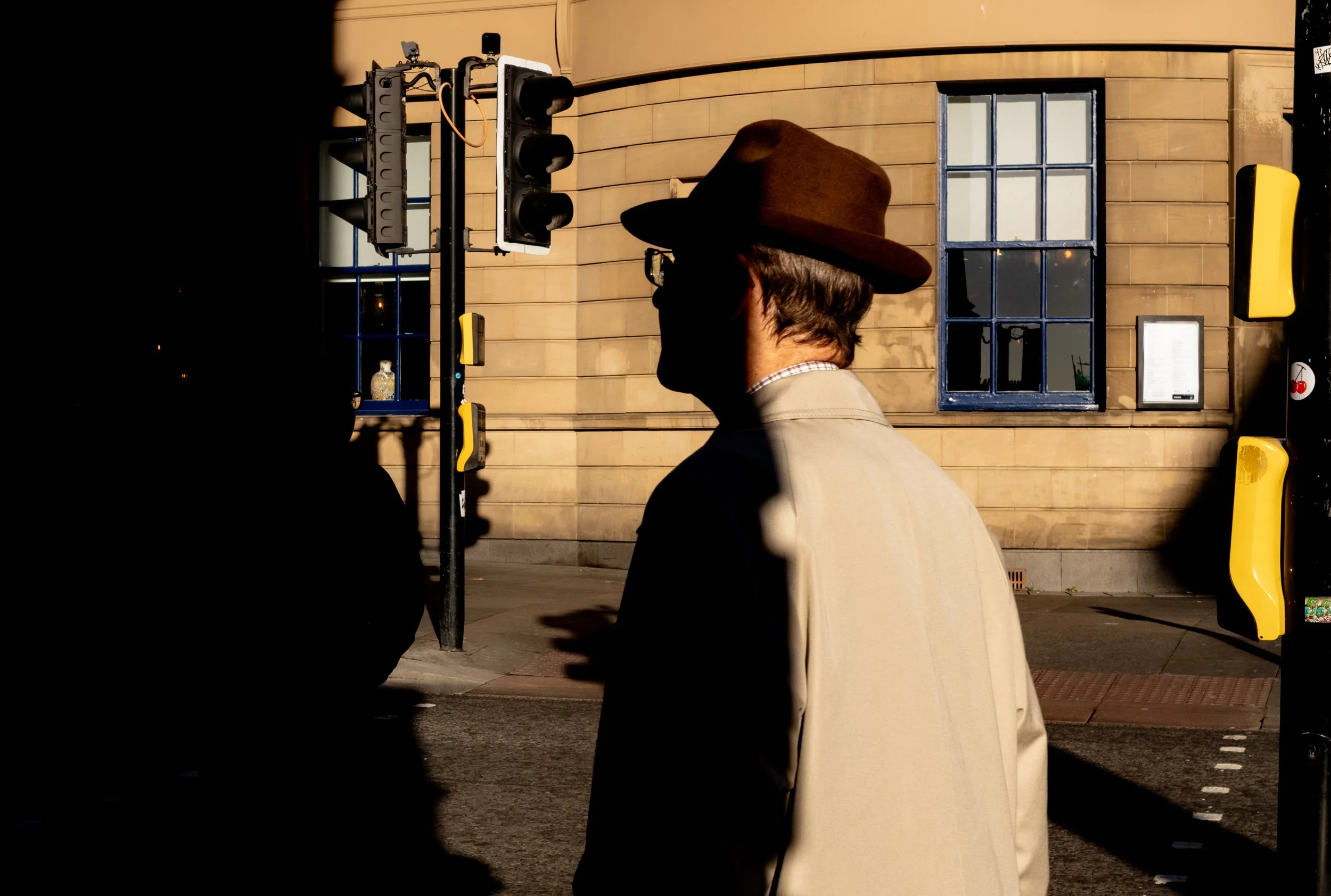 A person with gray hair wearing a brown wide-brimmed hat and glasses stands at a pedestrian crossing at night, with a beige building with a blue-framed window behind them. Traffic lights and yellow barriers are also visible.