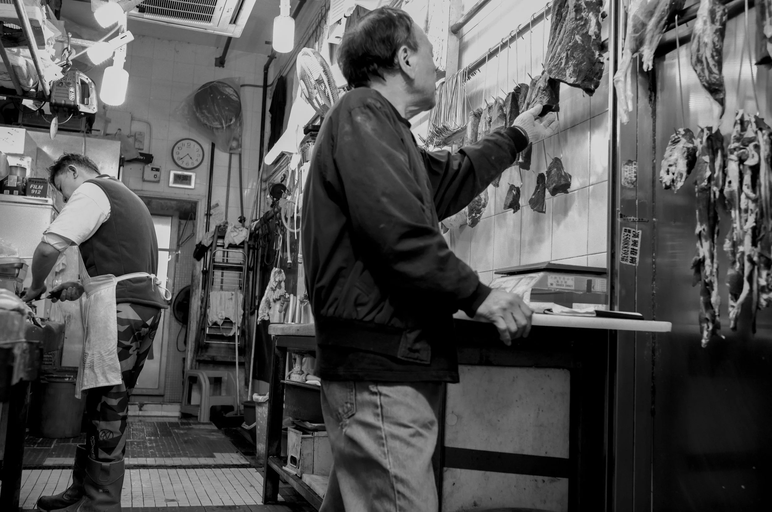 A man wearing gloves and a jacket working on hanging meat in a butcher shop, with another man preparing food in the background.