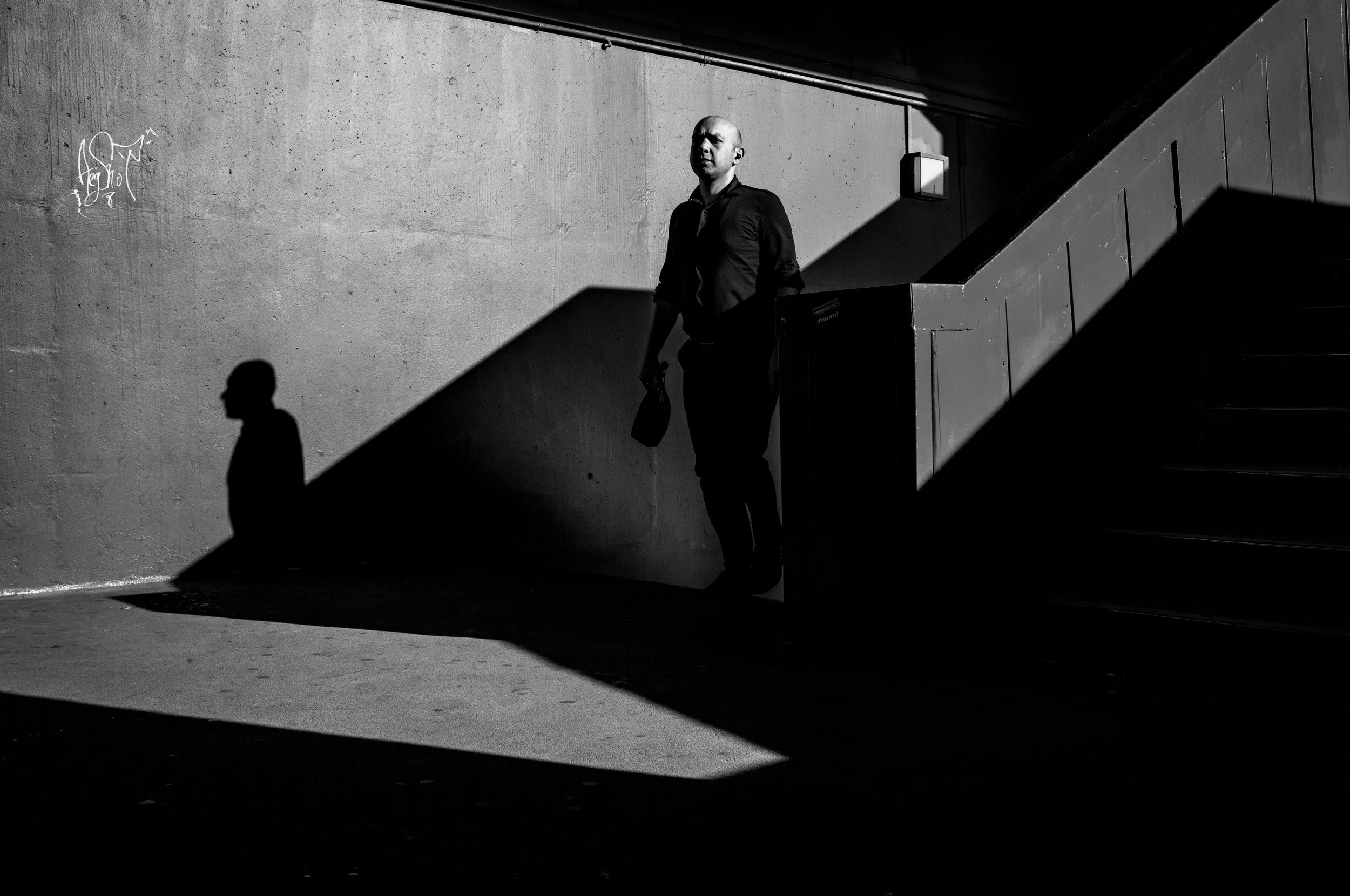 A man standing in a stairwell with strong shadows and contrast, illuminated partially by sunlight, creating sharp shadows on the wall and stairs.