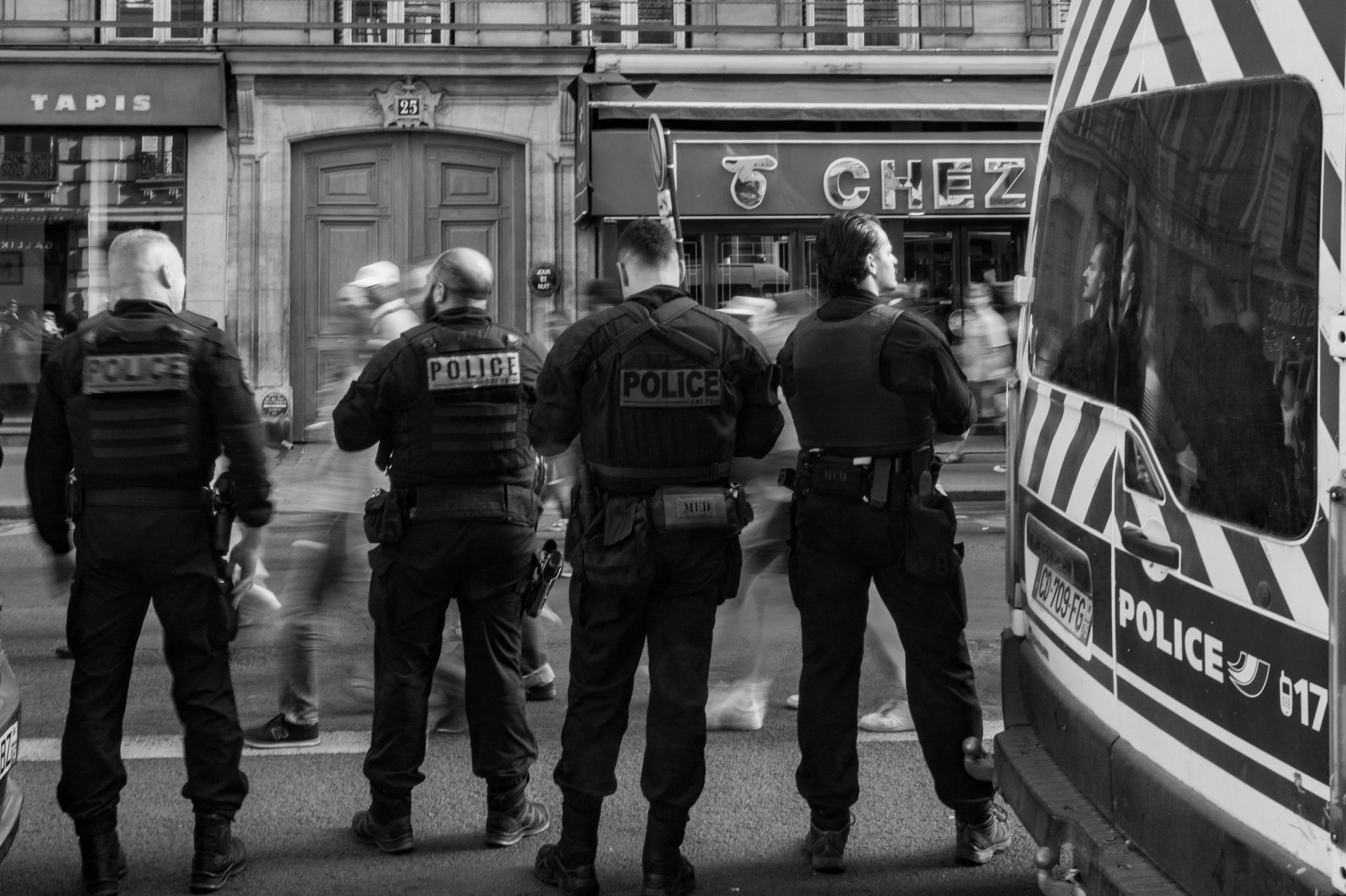 Four police officers standing on a city street near a police vehicle and a car with blurred pedestrians passing by.