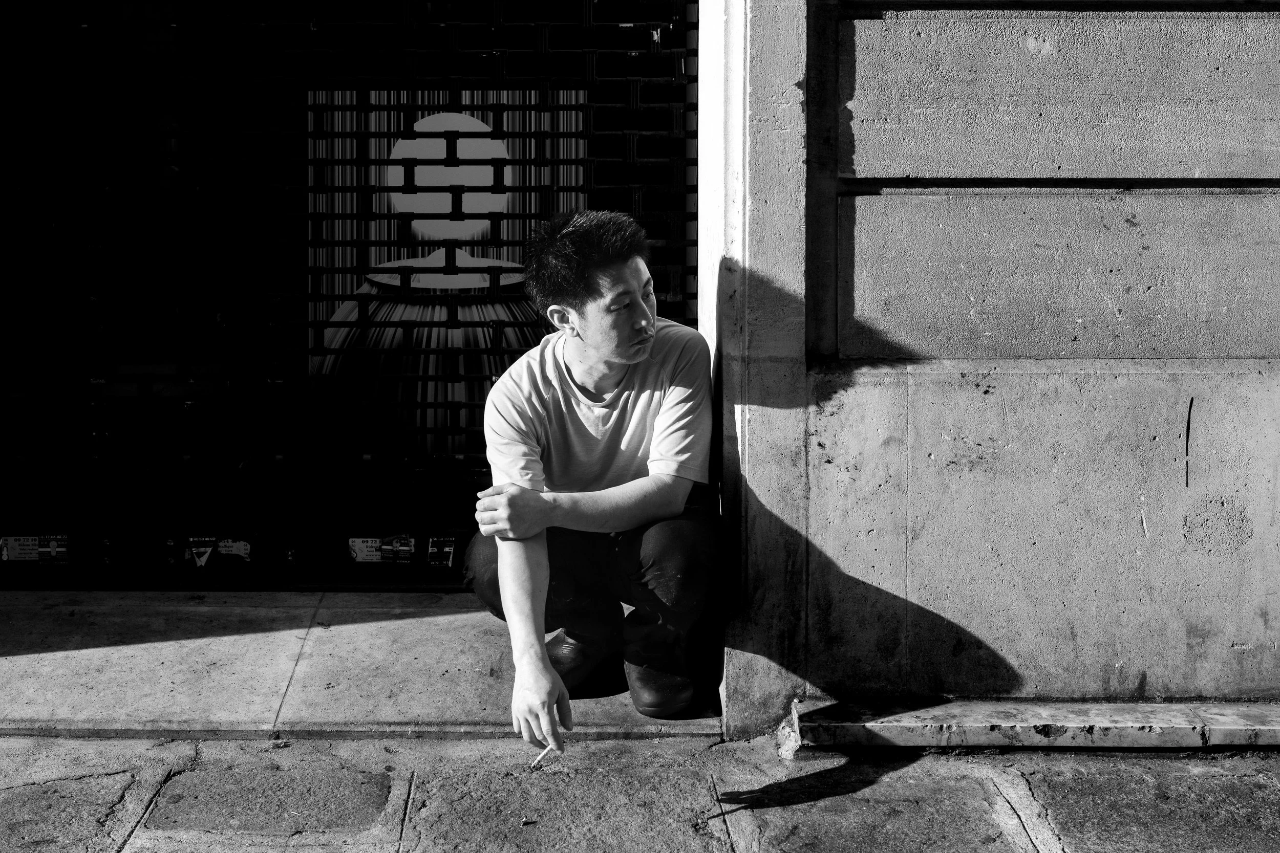 A young man with dark hair in casual clothing crouching outdoors against a concrete wall, with sunlight casting shadows. The background includes a metal gate with a circular design.