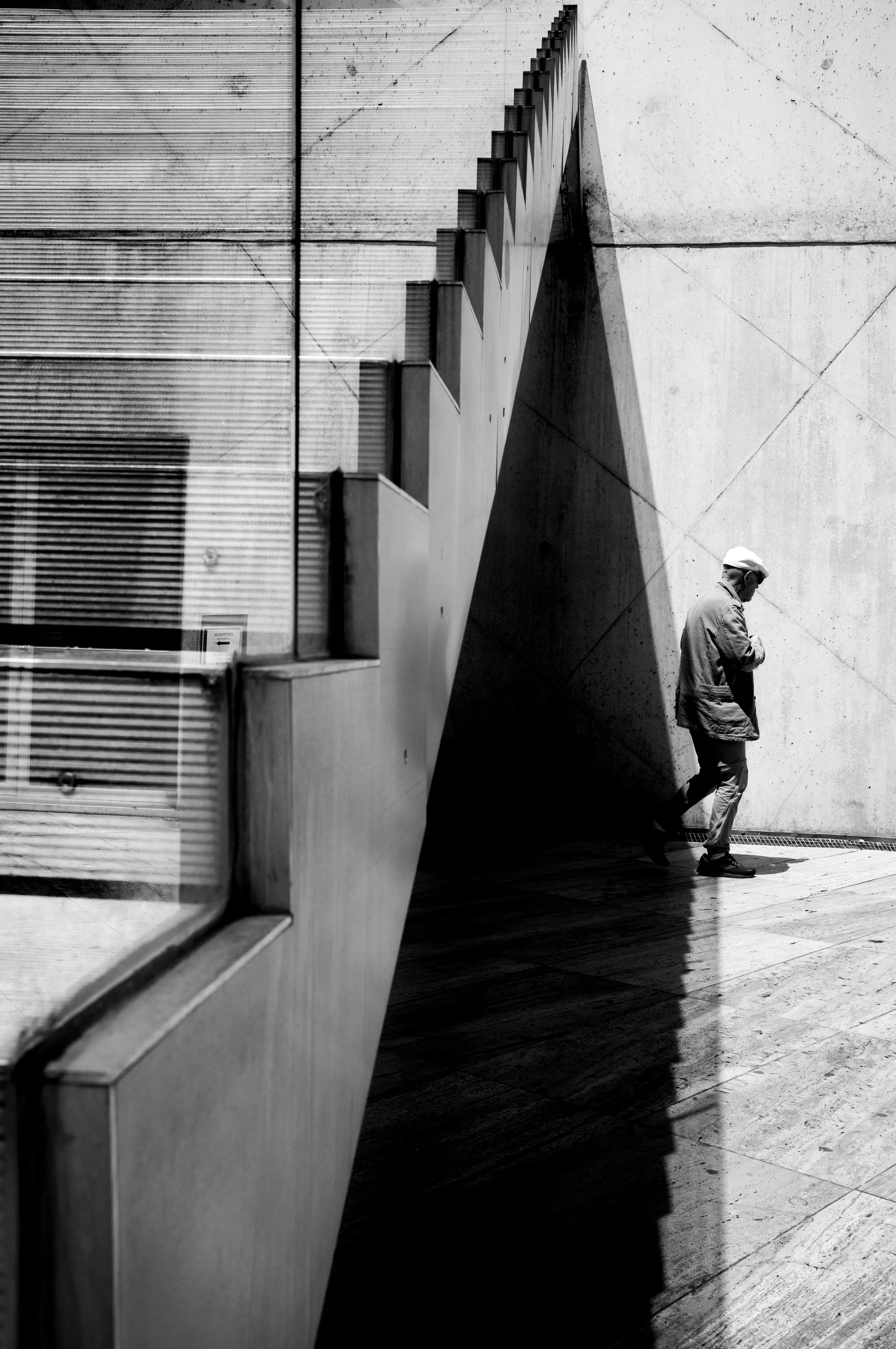 A man wearing a cap and jacket walks along a sidewalk next to a large concrete wall. Shadows are cast on the ground and wall, creating a stark contrast in the black and white photo. A glass window reflects part of the scene.