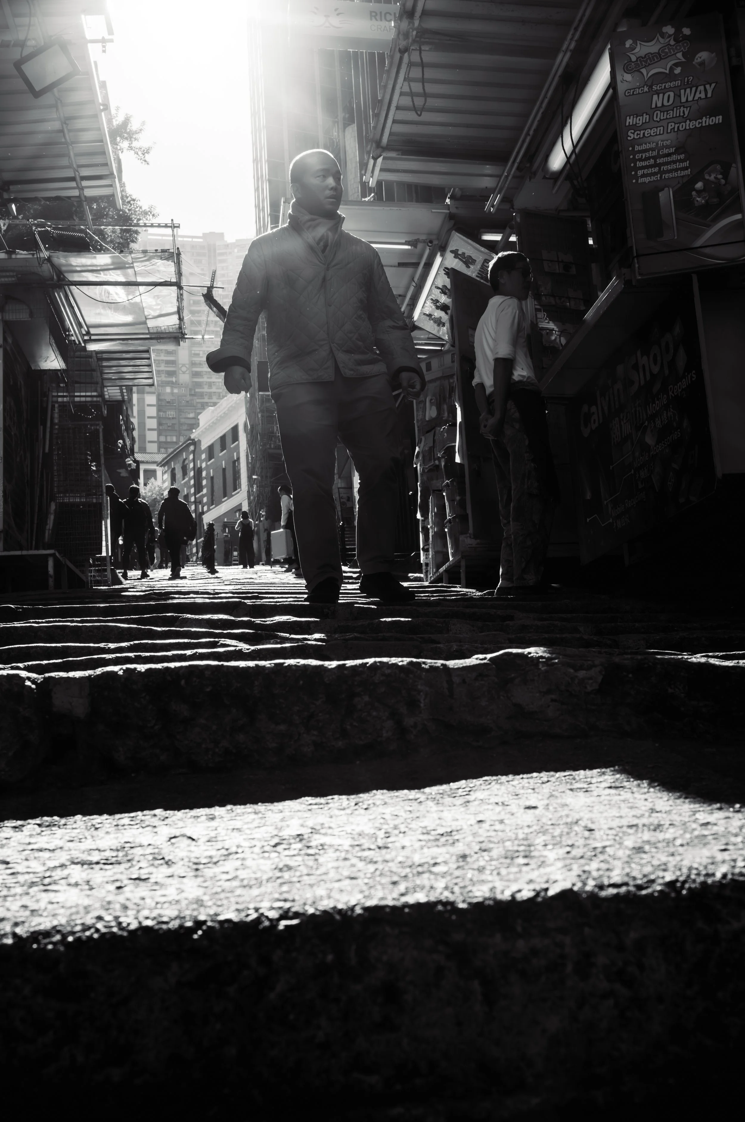 Black and white photo of a busy street scene with people walking and standing near shops. The sunlight creates bright highlights and shadows, with some buildings and signs visible.