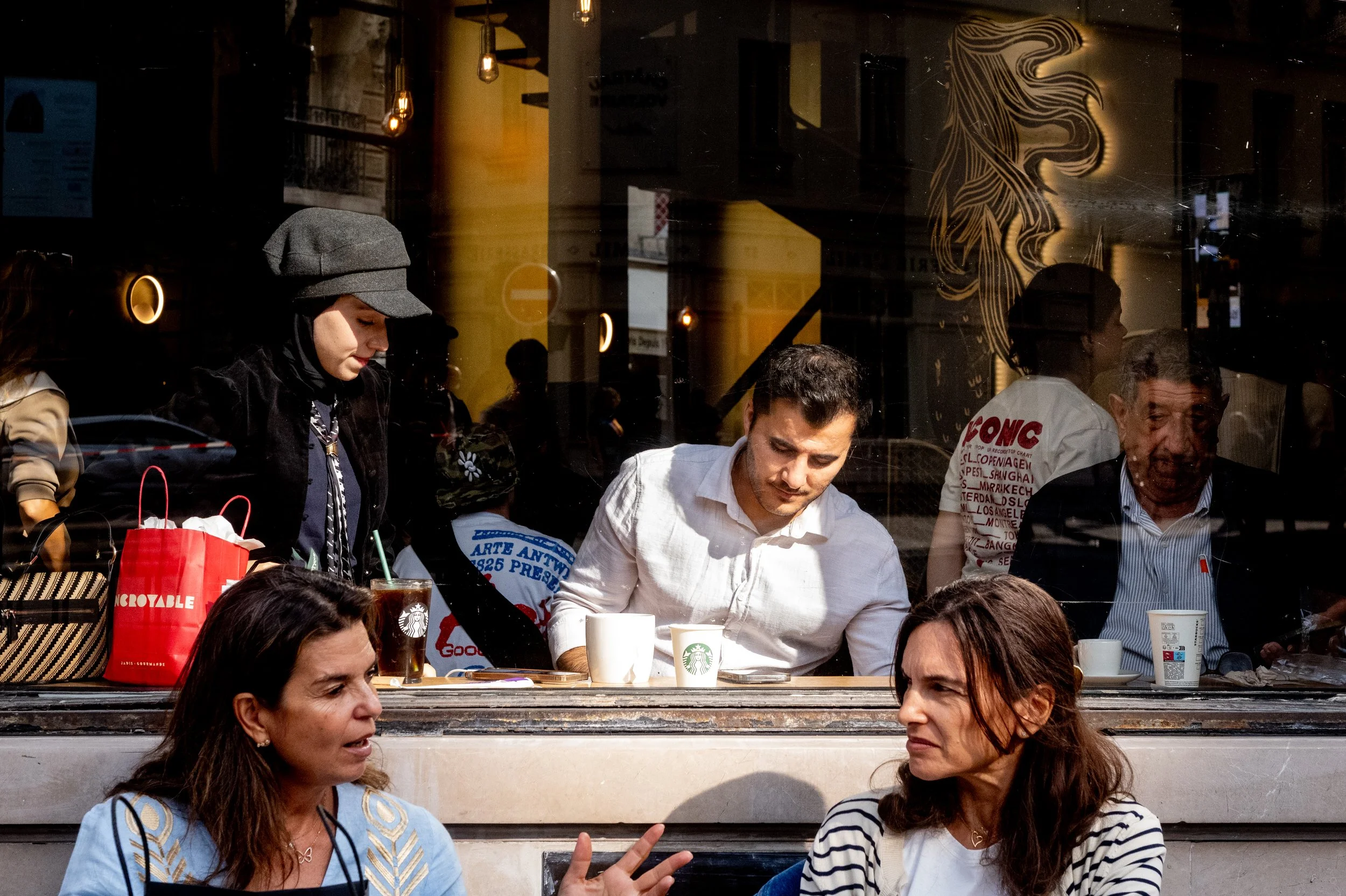 People sitting at a restaurant window, socializing and having drinks outside. The reflection shows street scenes and light fixtures inside the restaurant.