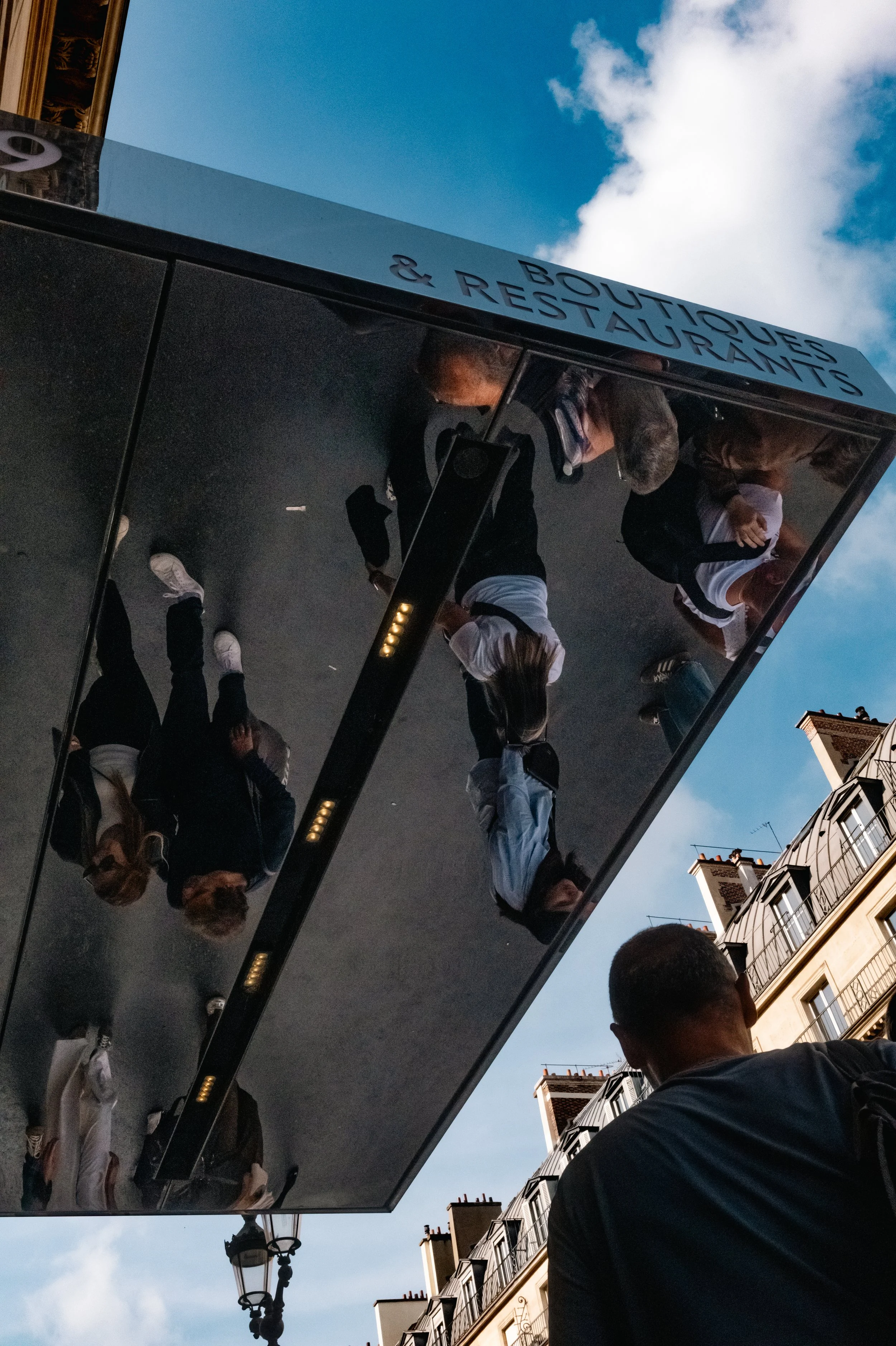 Reflection of people standing near a building and a street lamp in a large mirror on a building exterior, with a European-style building and blue sky with clouds in the background.