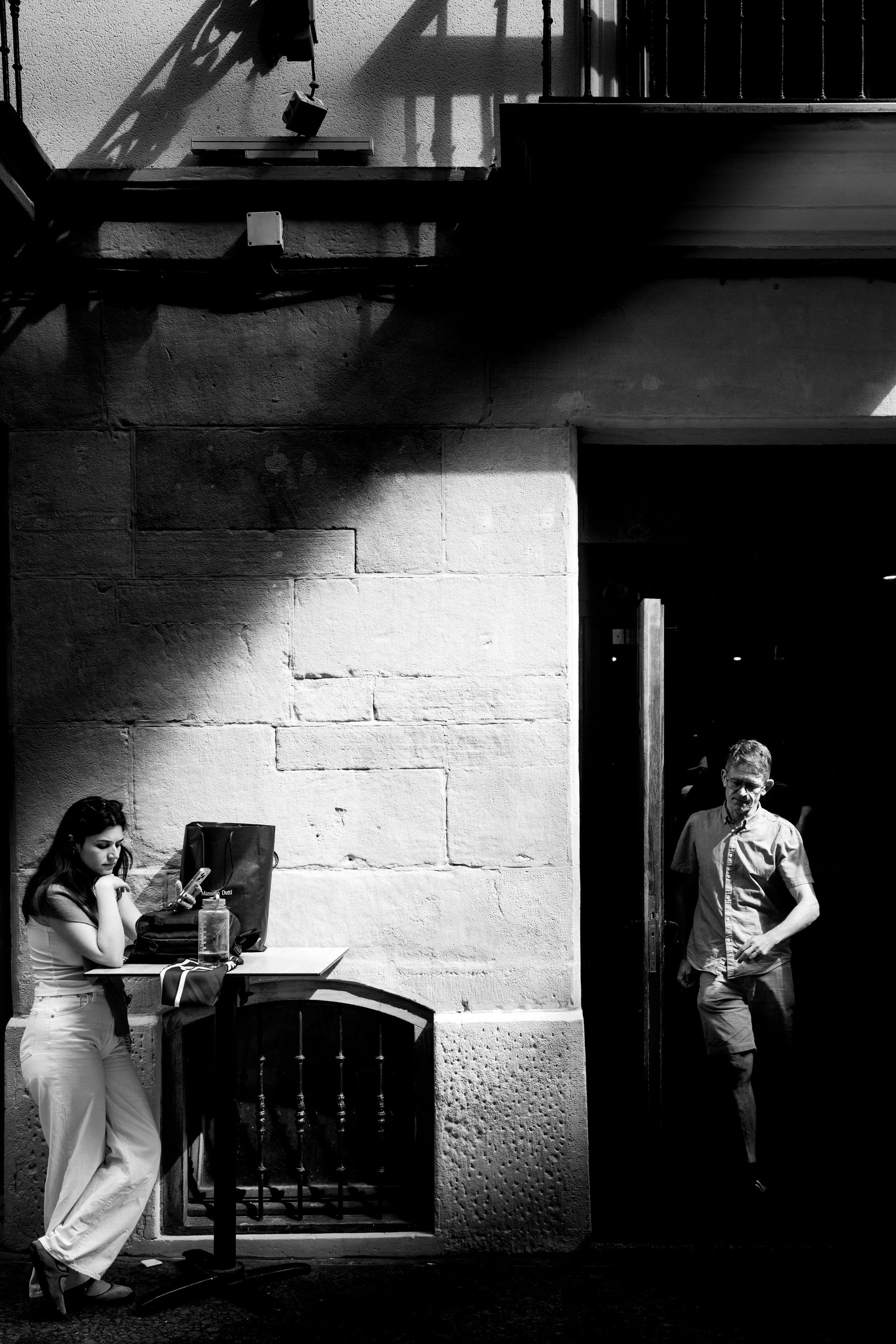 A black-and-white photo of a woman leaning against a table, looking at her phone, with a bag and water bottle in front of her. A man walks out of a building through a doorway, shadows cast on the wall behind them.