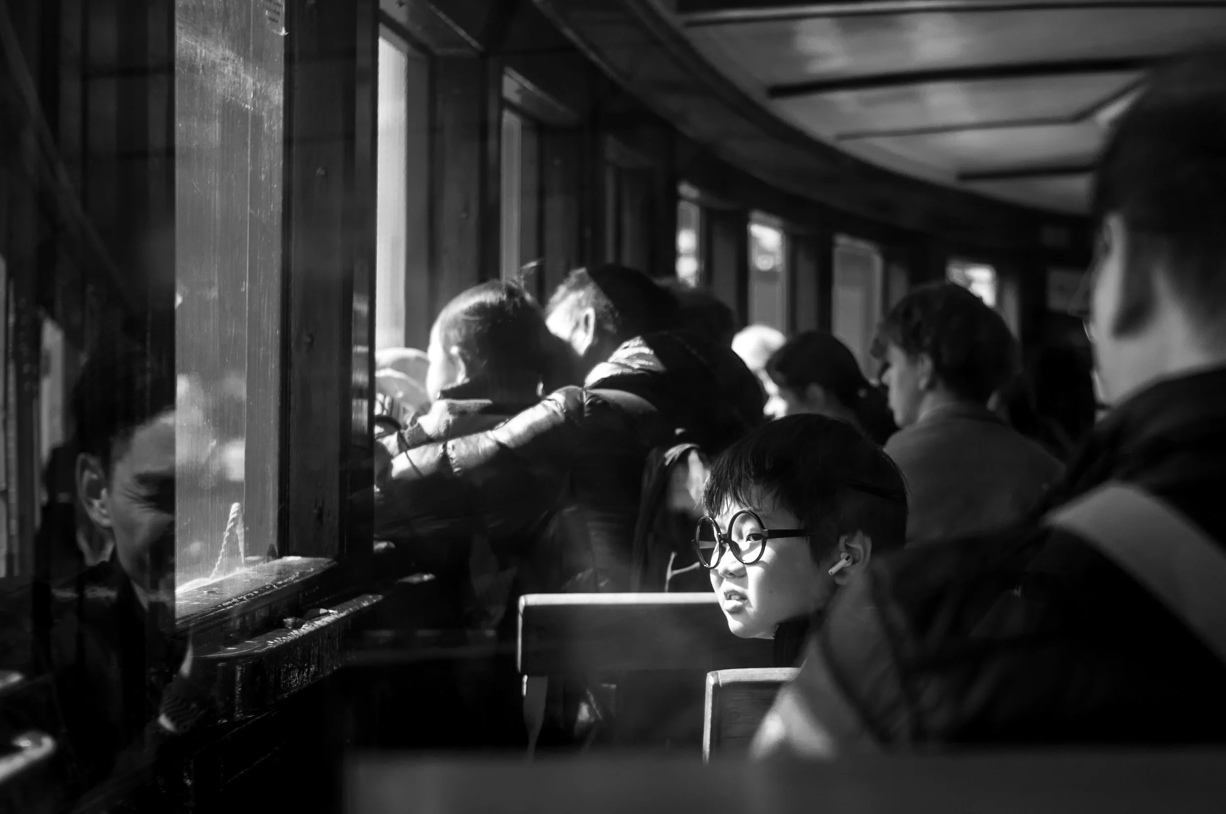 Black and white photo of several young people sitting closely together inside a train, near the window. They appear to be engaged in their own activities or conversations.