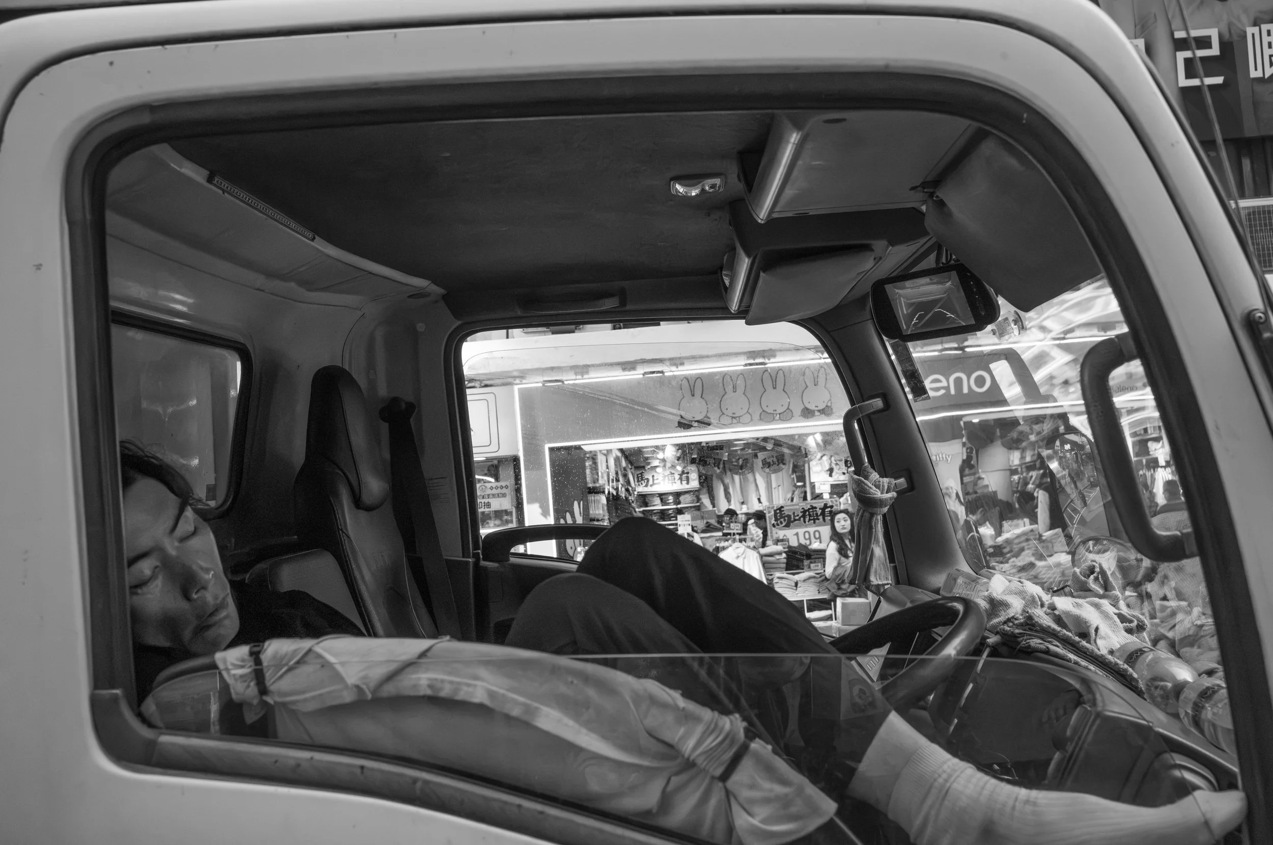 View from inside a vehicle showing a person sleeping or resting with their head on the window, and a market scene with bunny images and various products outside.