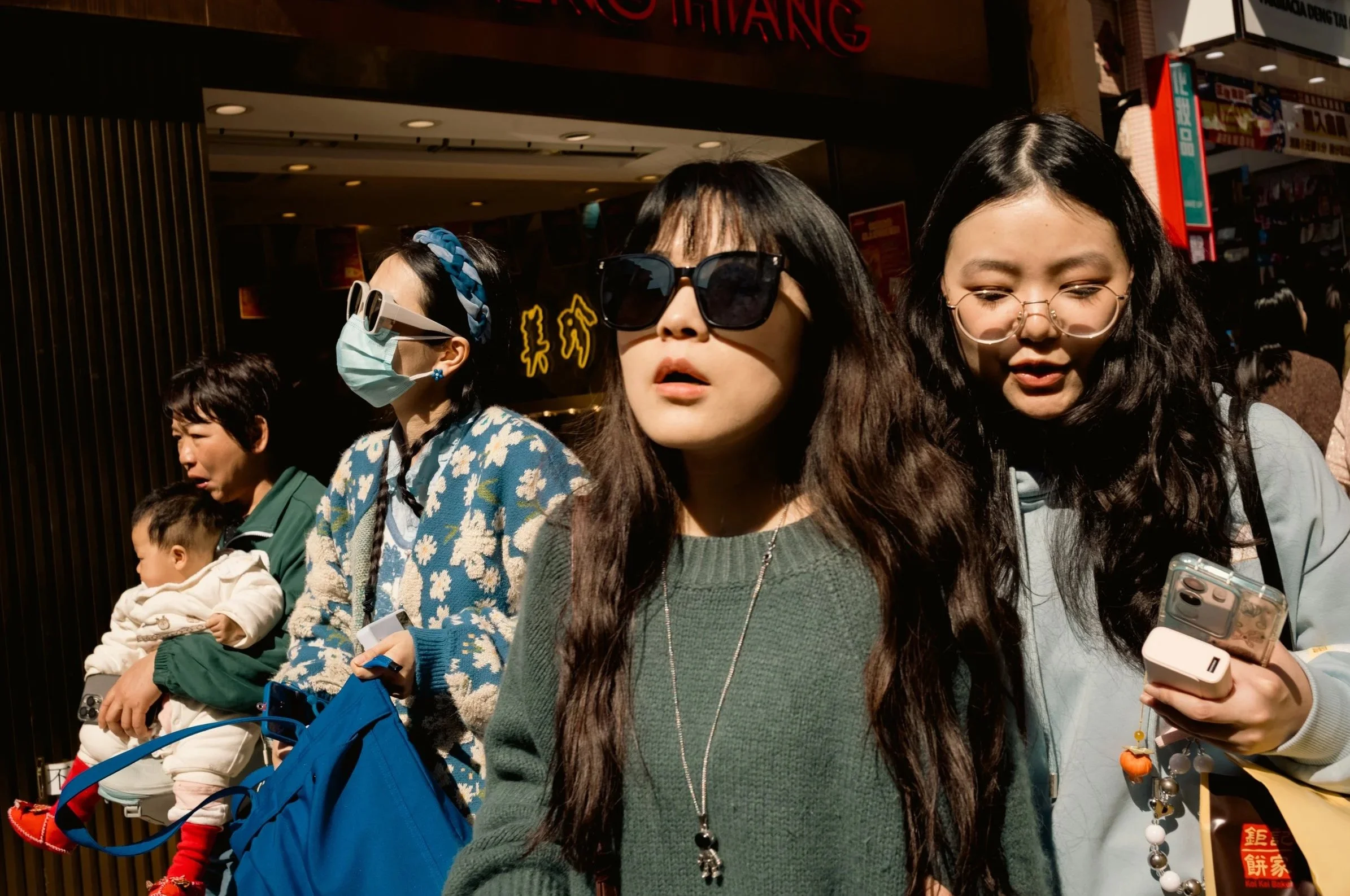 Group of women and children standing outside in sunny weather at a busy location, with storefronts and signs in the background.