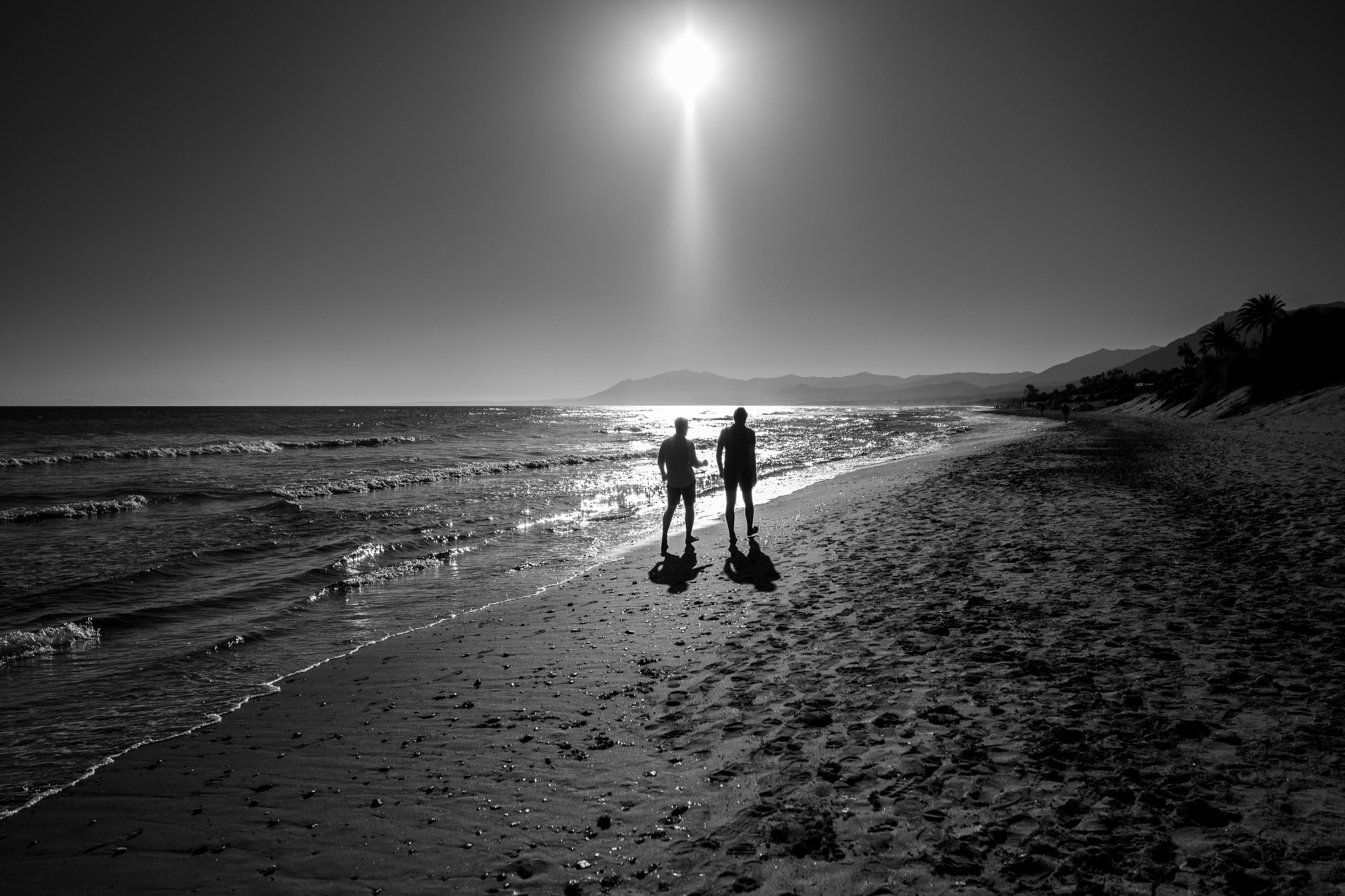 Two people walking along a beach at sunset or sunrise, with mountains and palm trees in the background, in black and white.