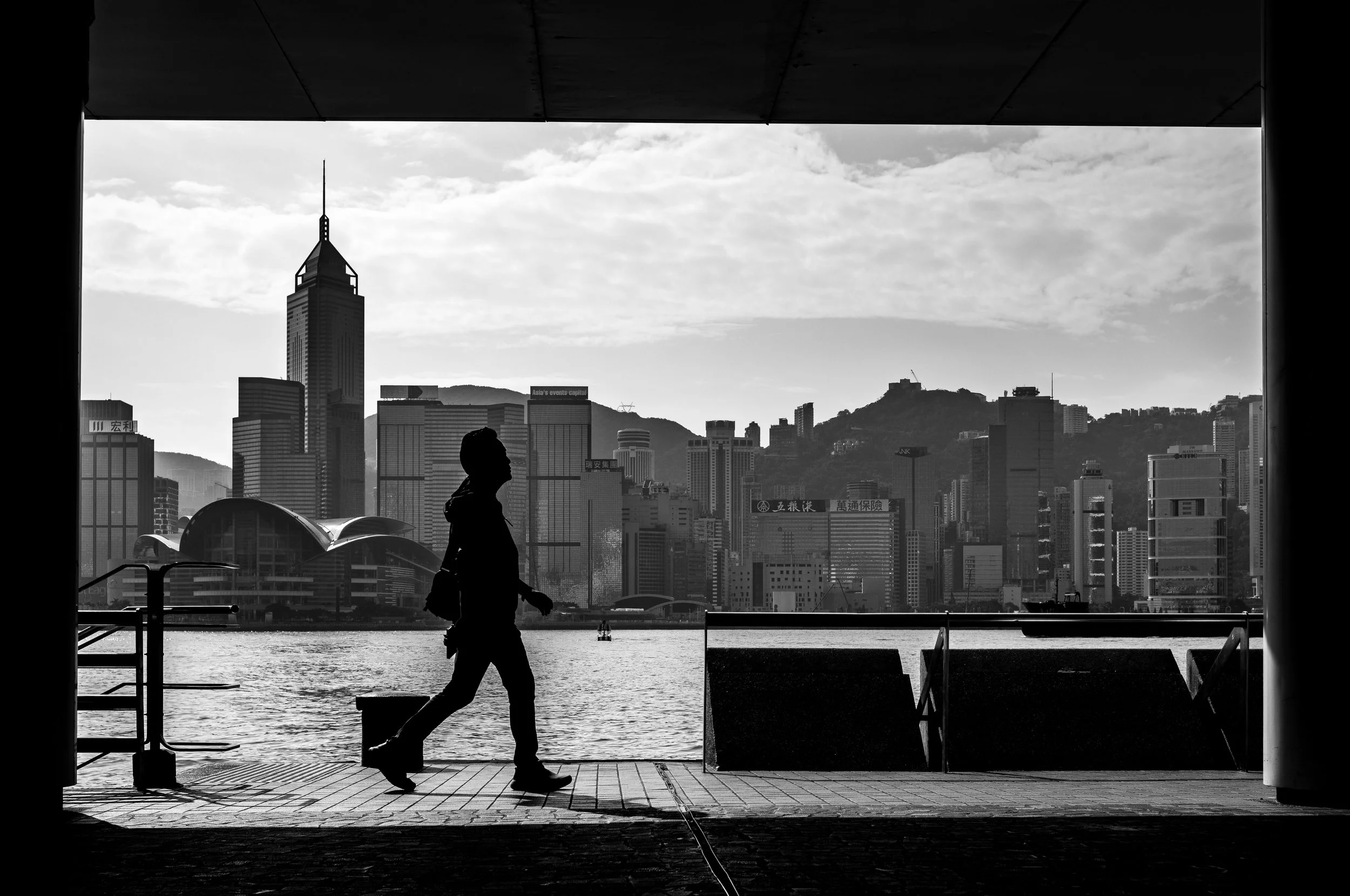 Silhouette of a person walking along a waterfront with a city skyline and mountains in the background, viewed from inside a building through a large window.