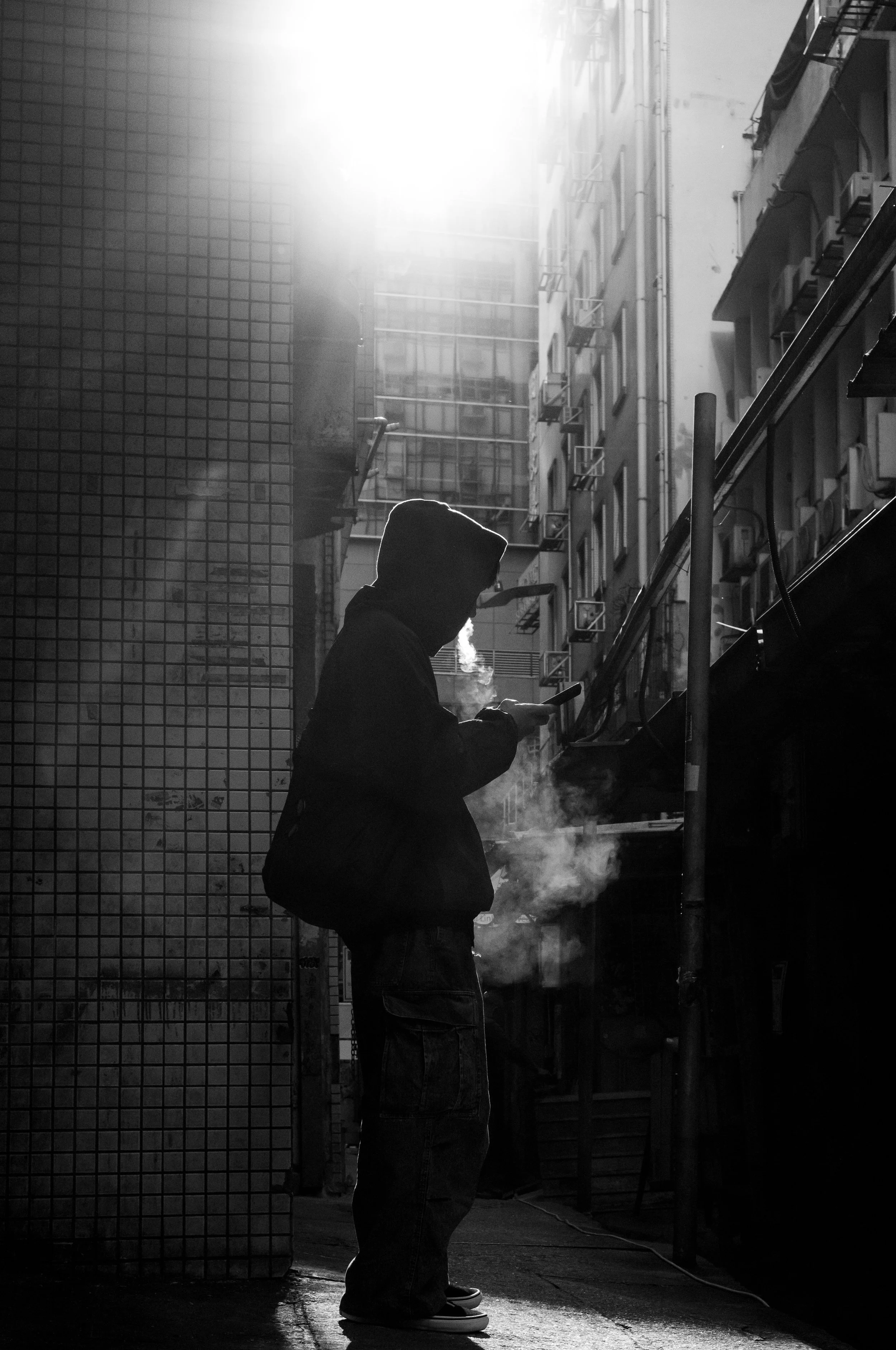 A person wearing a hoodie, standing on a city sidewalk, looking at a cellphone, with buildings and fire escapes in the background. The scene is in black and white with sunlight illuminating the scene.