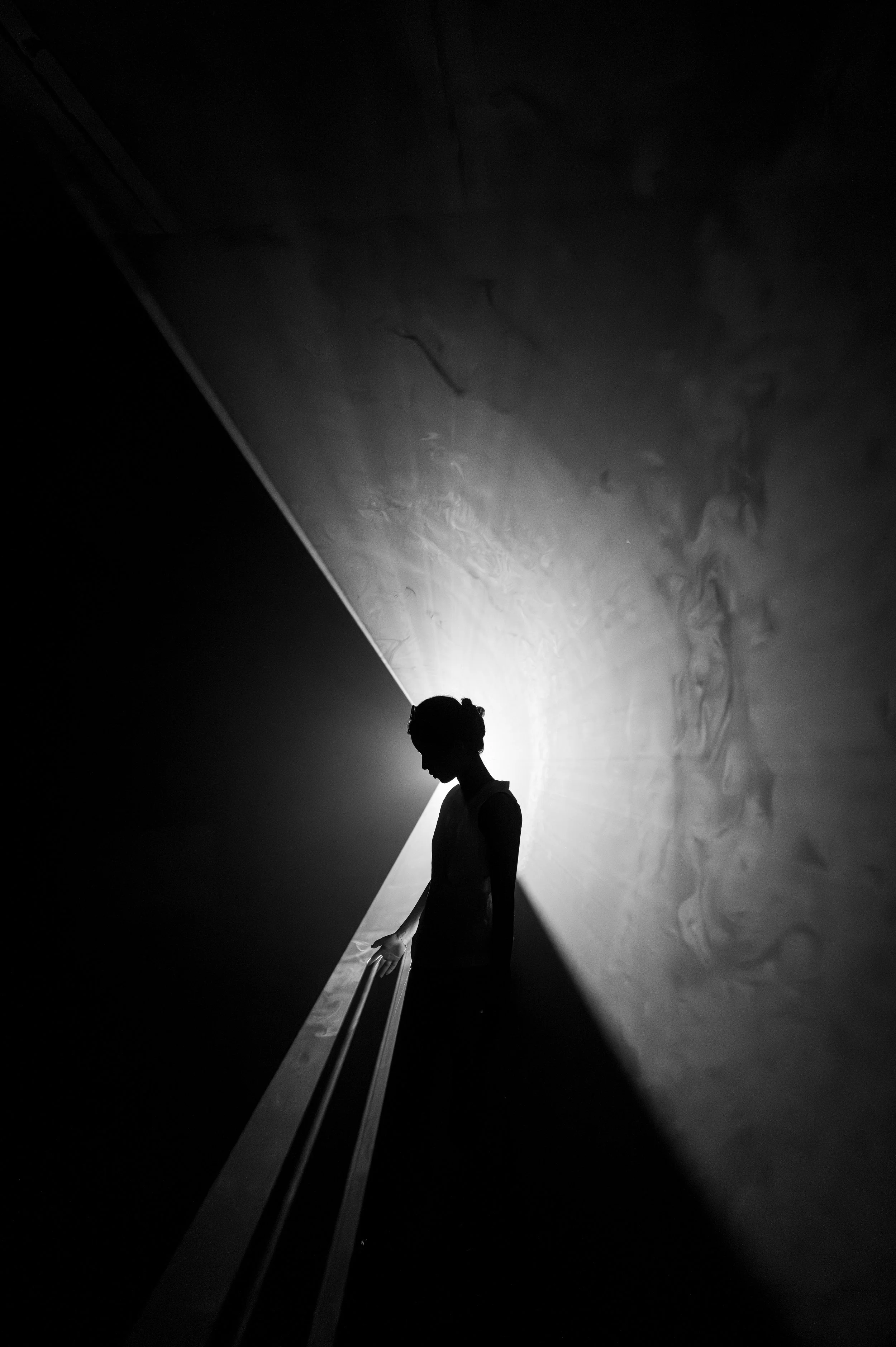A black and white photograph of a person standing on a staircase, with a dramatic, light-illuminated wall in the background. The person's silhouette is visible, and they are partially leaning on the staircase railing.