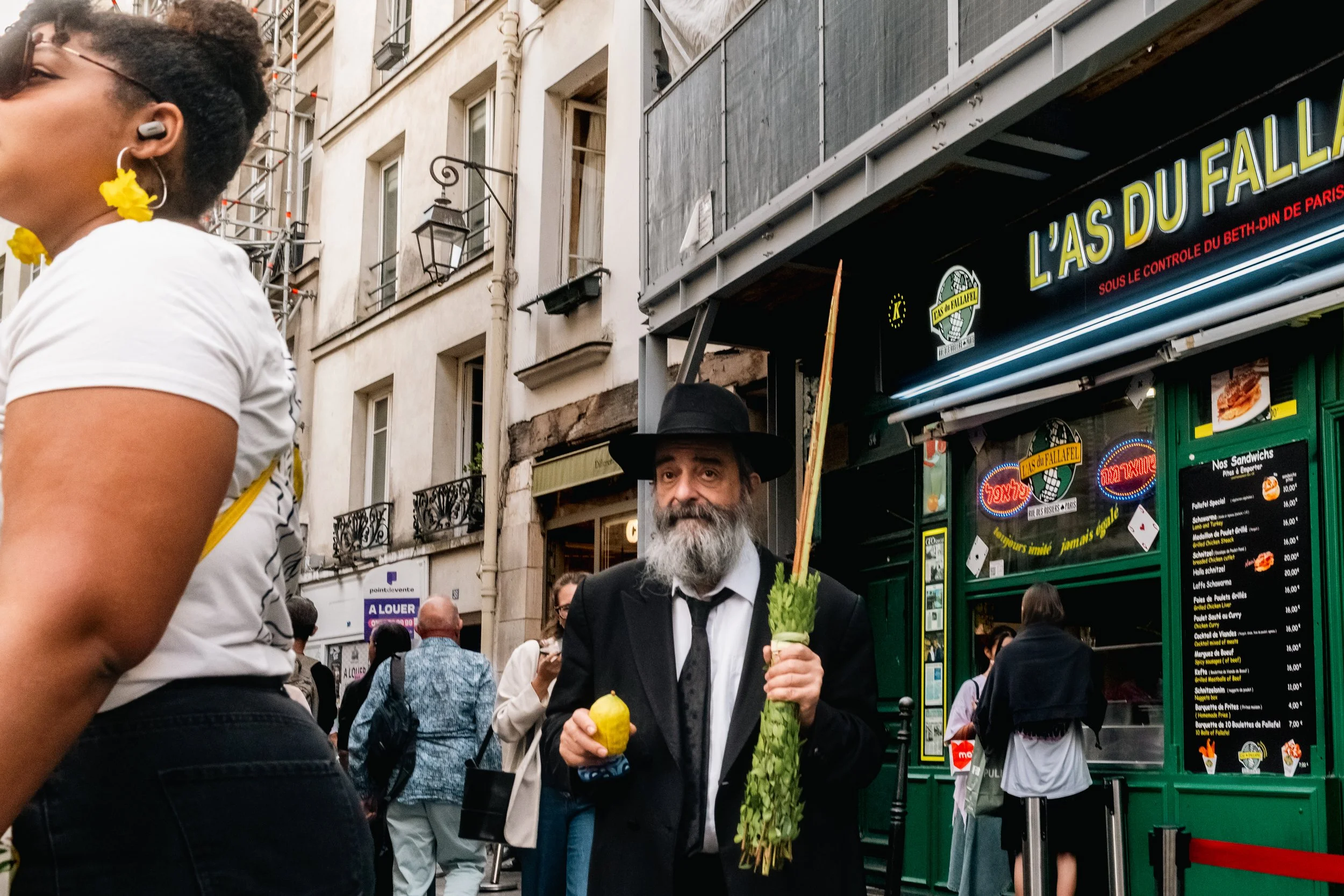 A man dressed in a black suit, white shirt, and black hat is holding two large vegetables, a pineapple and a celery stalk, in front of a busy street scene in Paris. There are several people around him, and storefronts with signs, including a neon sig