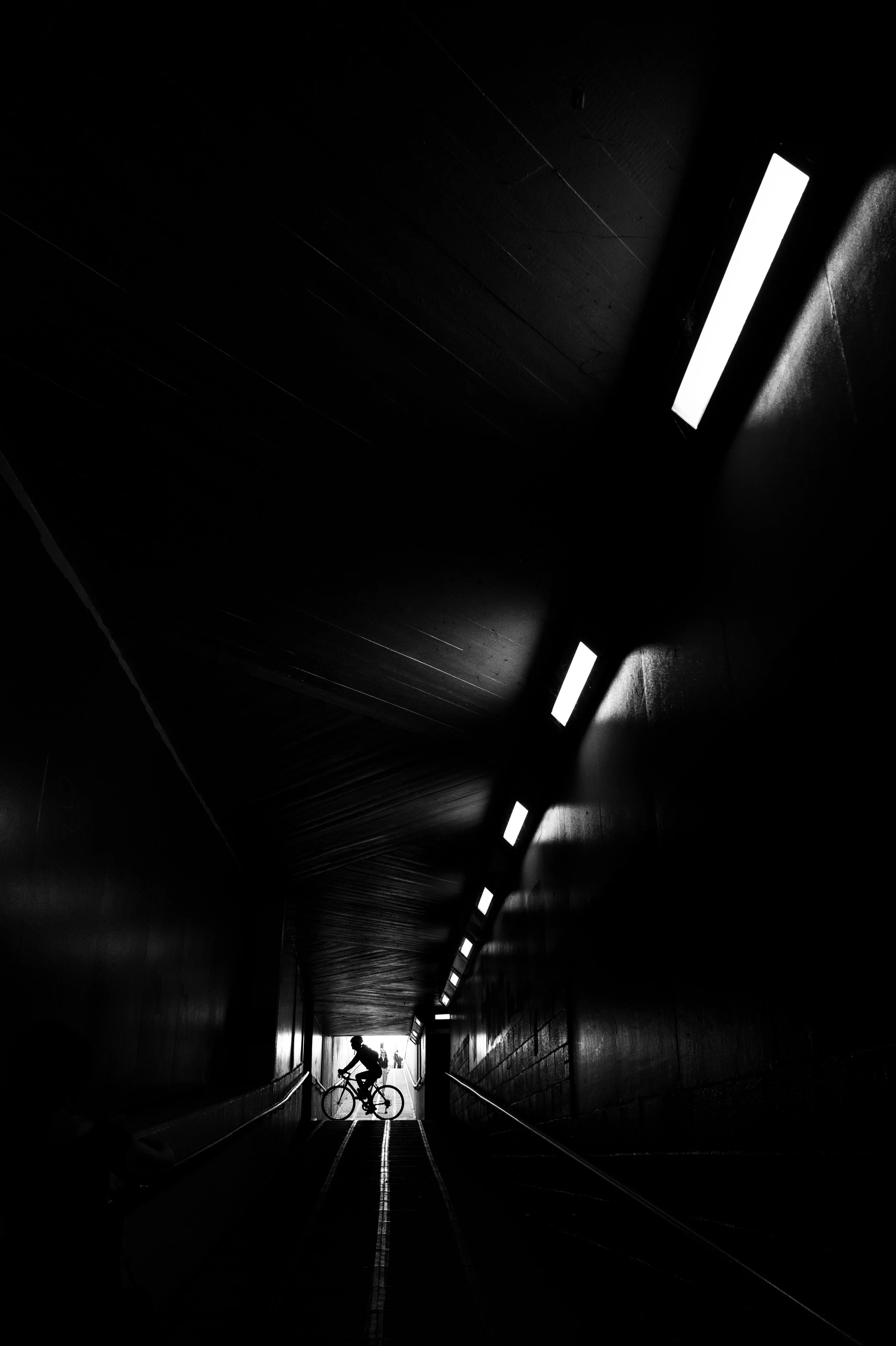 Silhouette of a person riding a bicycle in a dark tunnel with bright rectangular lights on the ceiling and a person at the end of the tunnel in the distance.