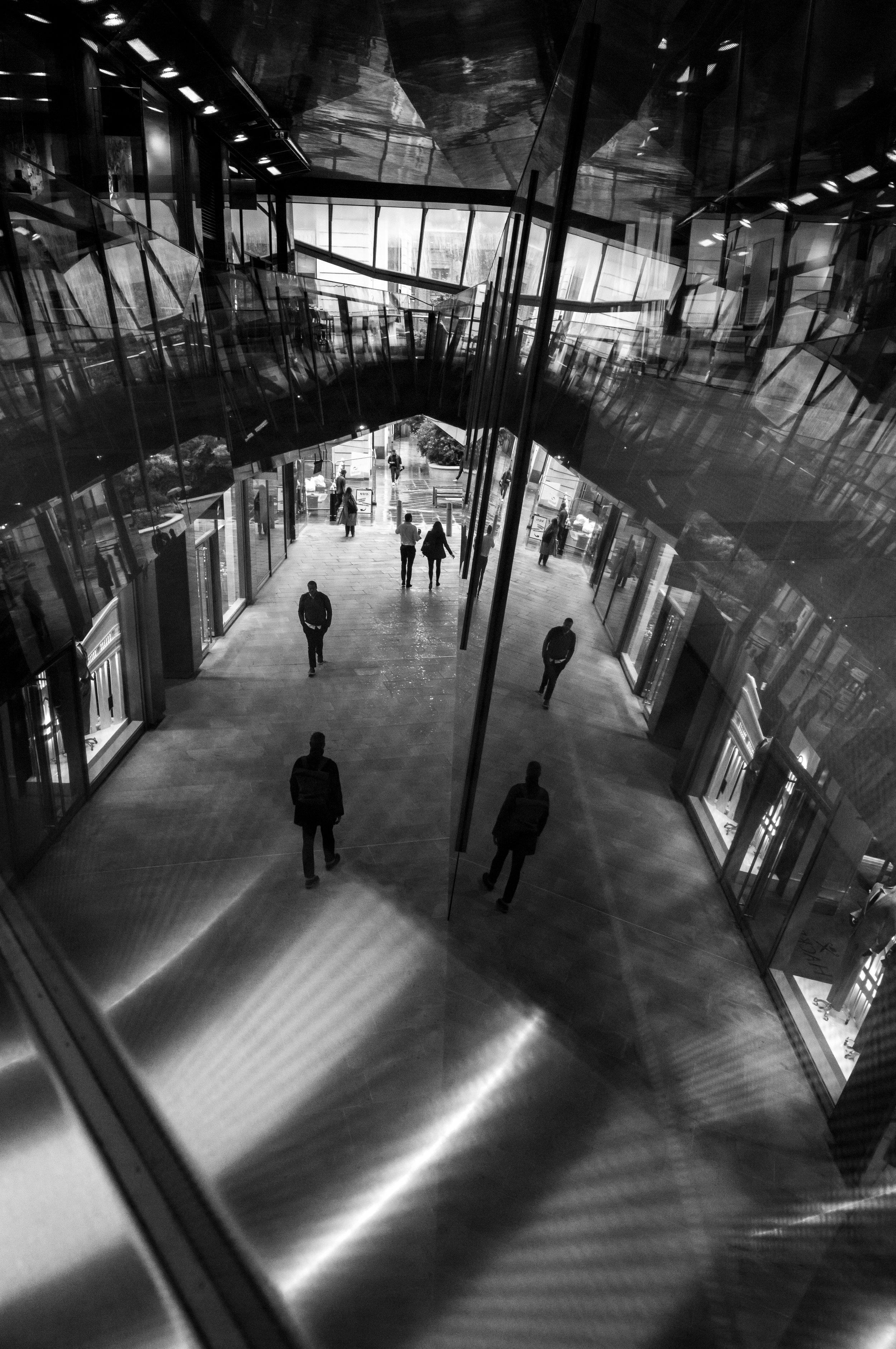 A black and white photo of an indoor shopping mall. The perspective is from inside an elevator looking out through the glass walls. Several people are walking on the mall's corridor, and there are multiple shops visible on either side.