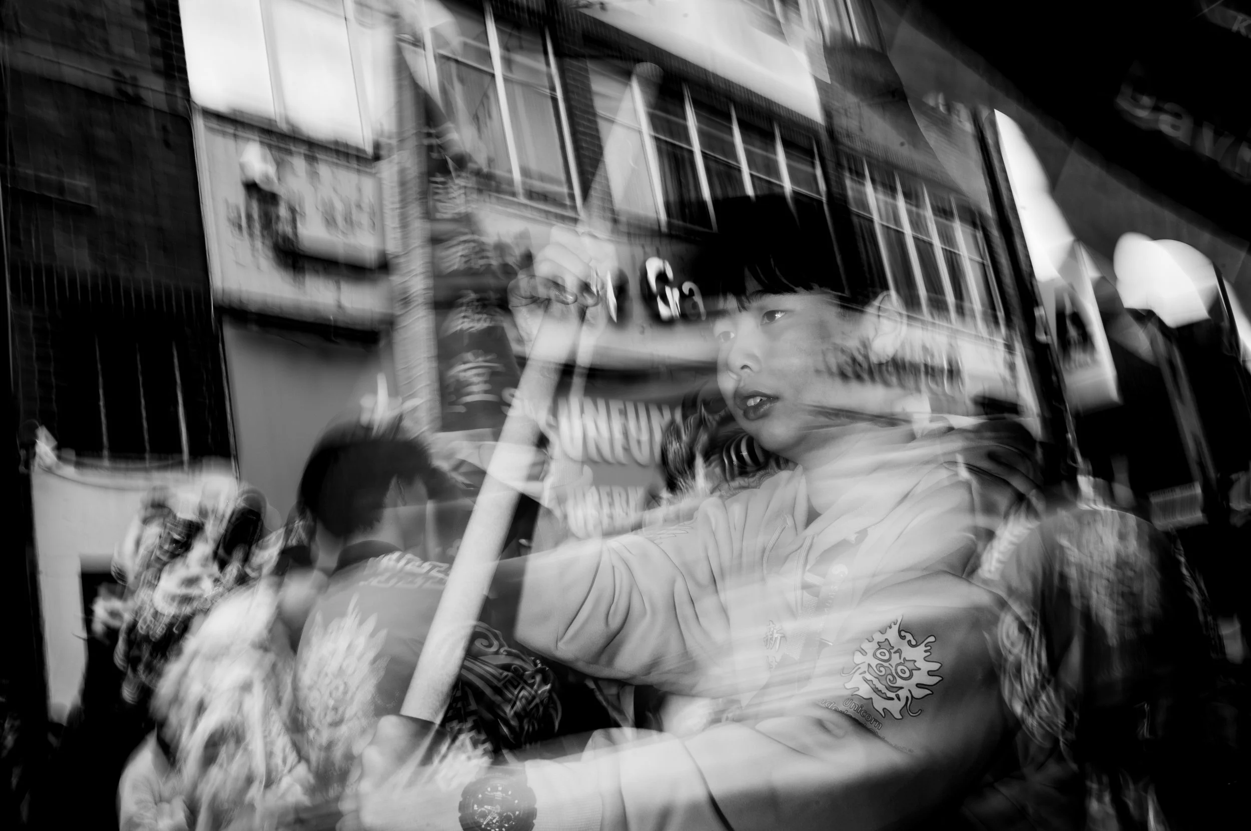 A black and white photo captures a young boy sitting in a vehicle, with reflections of city buildings and signs visible on the window.