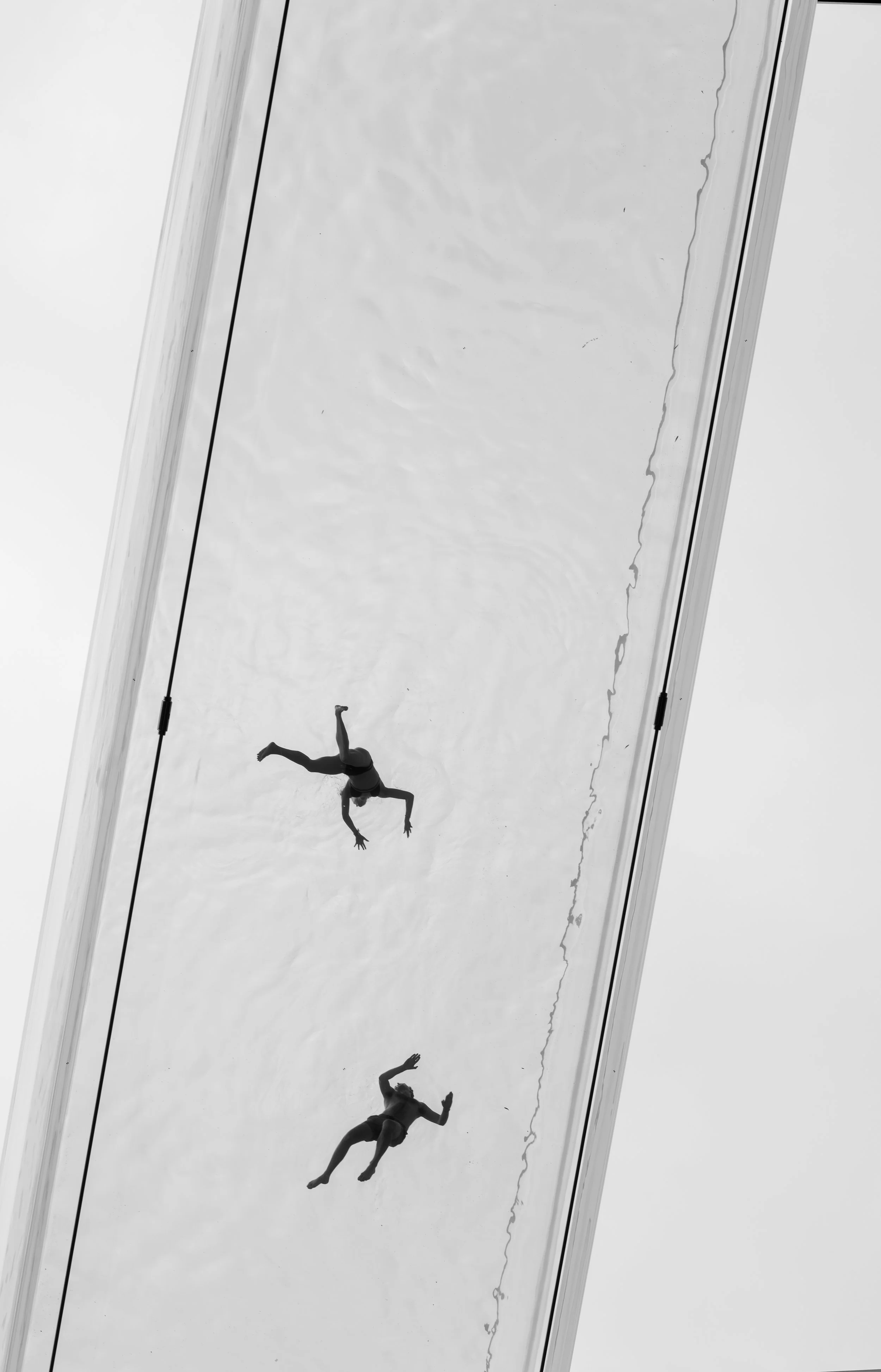 A person performing a dance or acrobatic move on a large mirror ceiling reflects their image upside down. The person is in mid-performance, wearing dark clothing, with one arm extended and one leg bent, against a clean, white background.