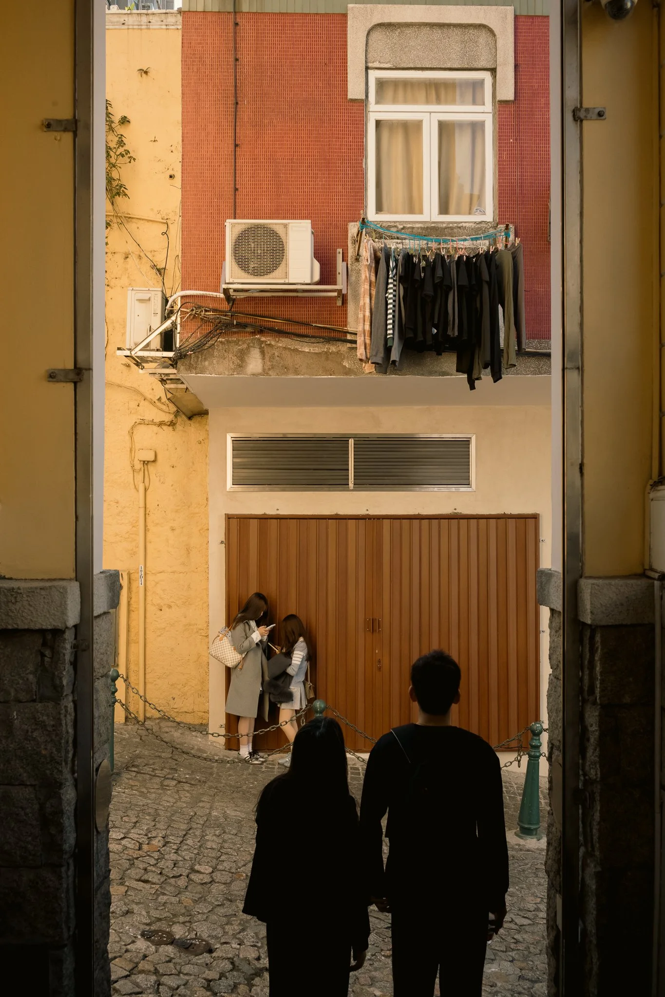 View through a stone archway showing two girls and two boys standing on a cobblestone street, with the girls talking to each other and the boys facing away, against a backdrop of closed brown door, yellow and red buildings, a window with beige curtai