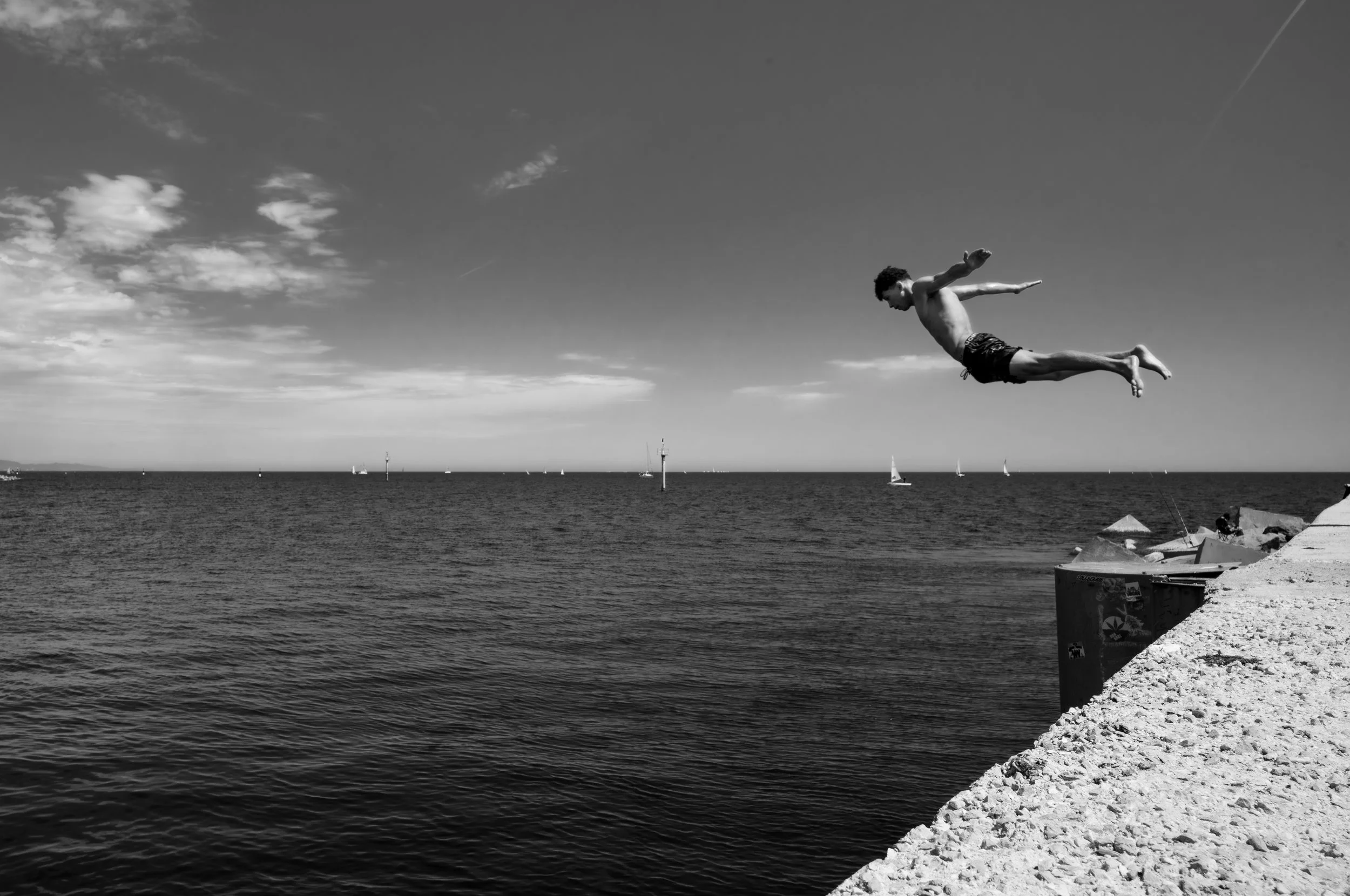 A young man is mid-air jumping off a rocky pier into the ocean on a clear day, with sailboats visible on the water and a partly cloudy sky in the background.