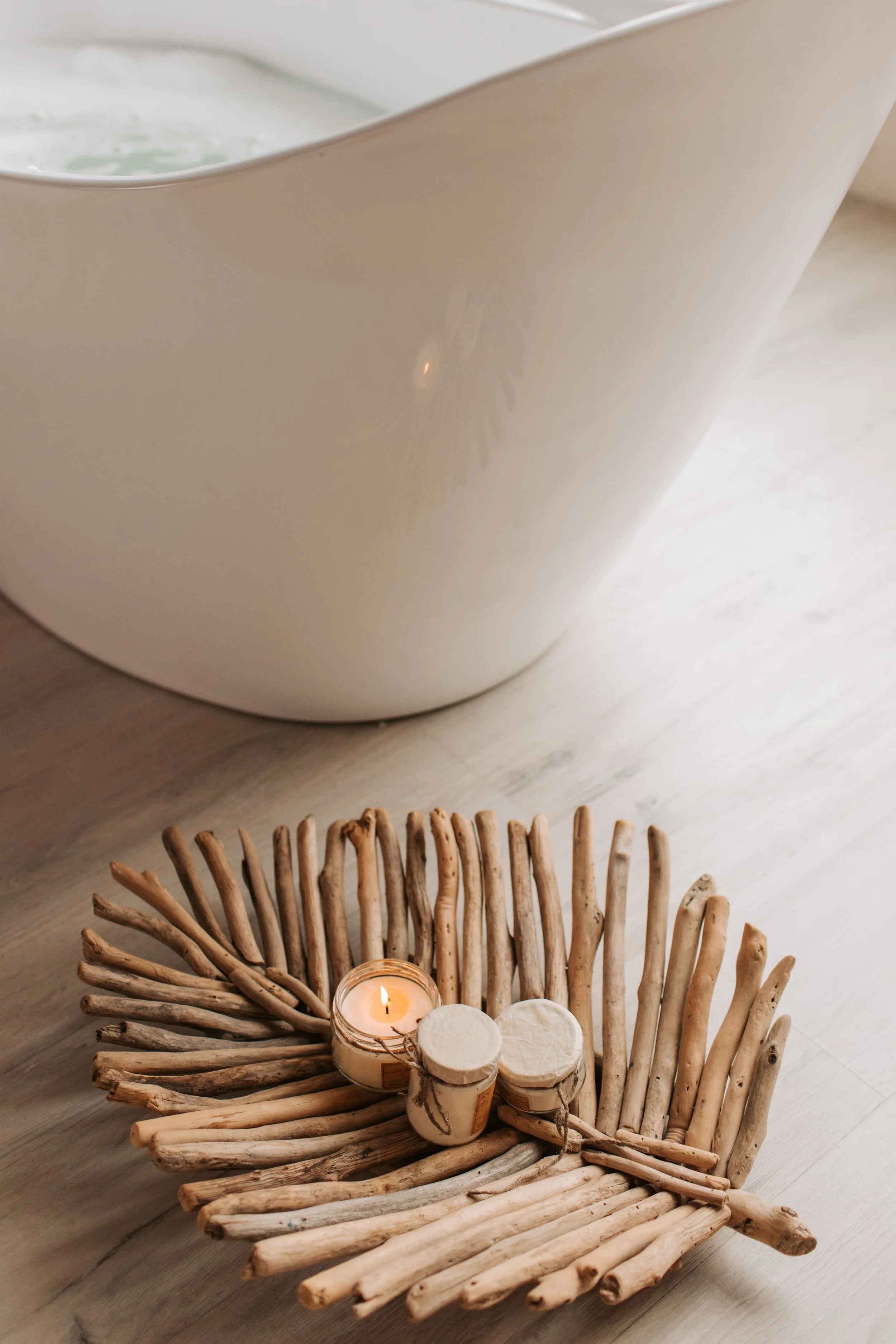 Decorative arrangement on a wooden surface featuring driftwood border, white candles, and a lit candle in a glass jar with twine.