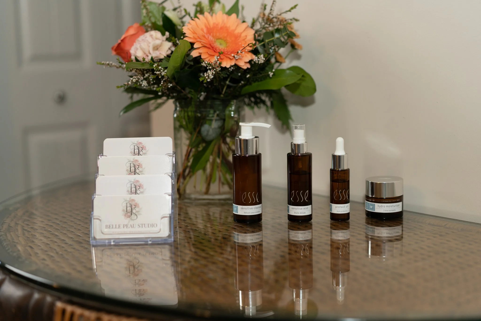 A bouquet of mixed flowers in a glass vase, placed on a table, accompanied by skincare products including bottles and a small jar, with a card holder displaying cards reading Belle Peau Studio.