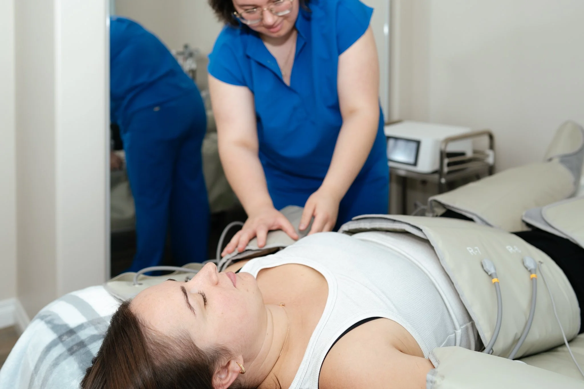 A woman lies on a hospital bed with her eyes closed, connected to medical wires and monitors. A medical professional in blue scrubs is attending to her, preparing her for a procedure in a clinical setting.