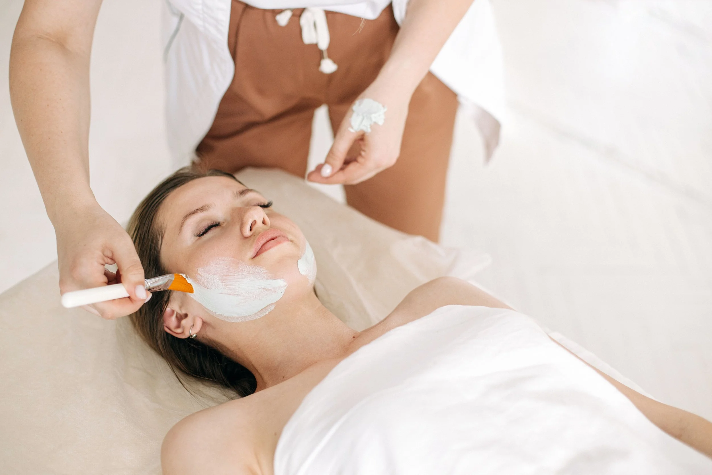 A woman lying on a bed getting a facial treatment, with a skincare mask being applied on her face by a professional.