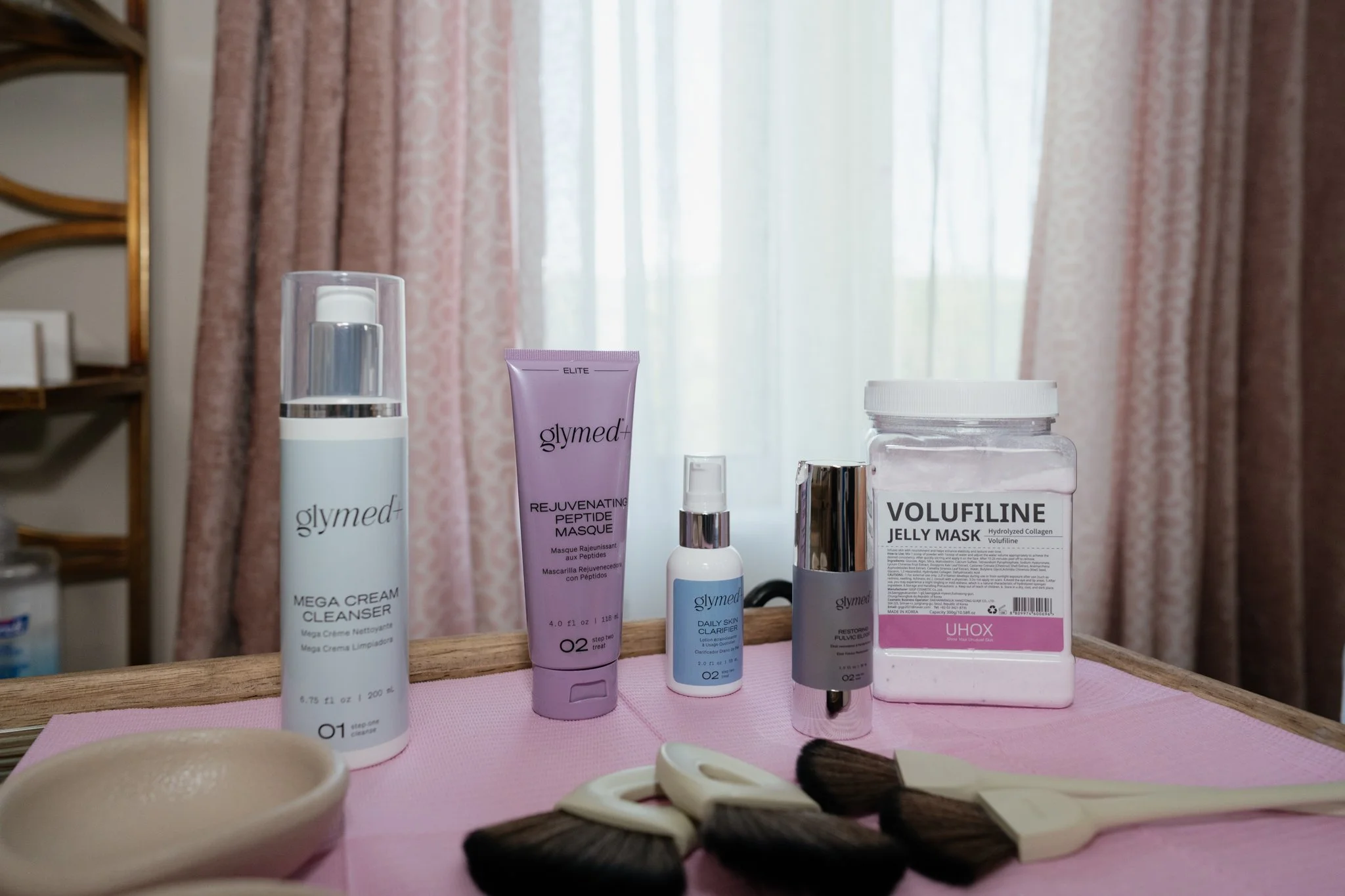 A collection of skincare and beauty products on a pink surface with a blurred background of a window with lace and pink curtains.