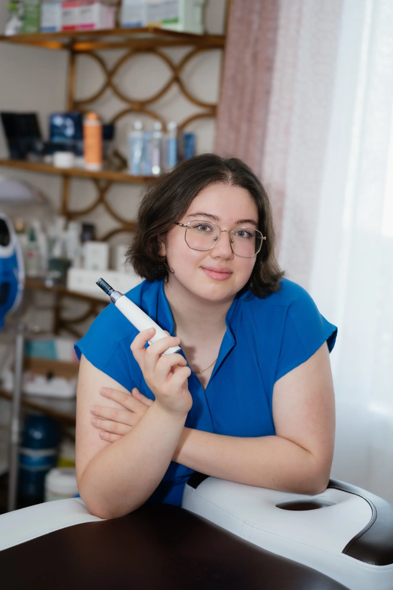 A young woman with glasses and shoulder-length brown hair, wearing a blue top, is sitting at a medical or physical therapy table, holding a pen, with an indoor setting that includes shelves with bottles and medical supplies in the background.