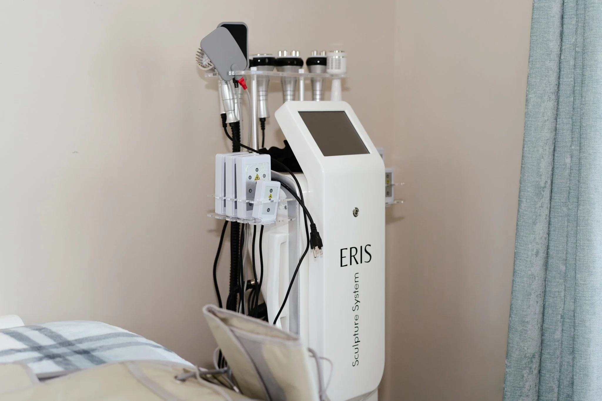 Medical equipment with a touchscreen monitor, placed in a hospital room near a bed with striped bedding, and curtains on the right side of the image.