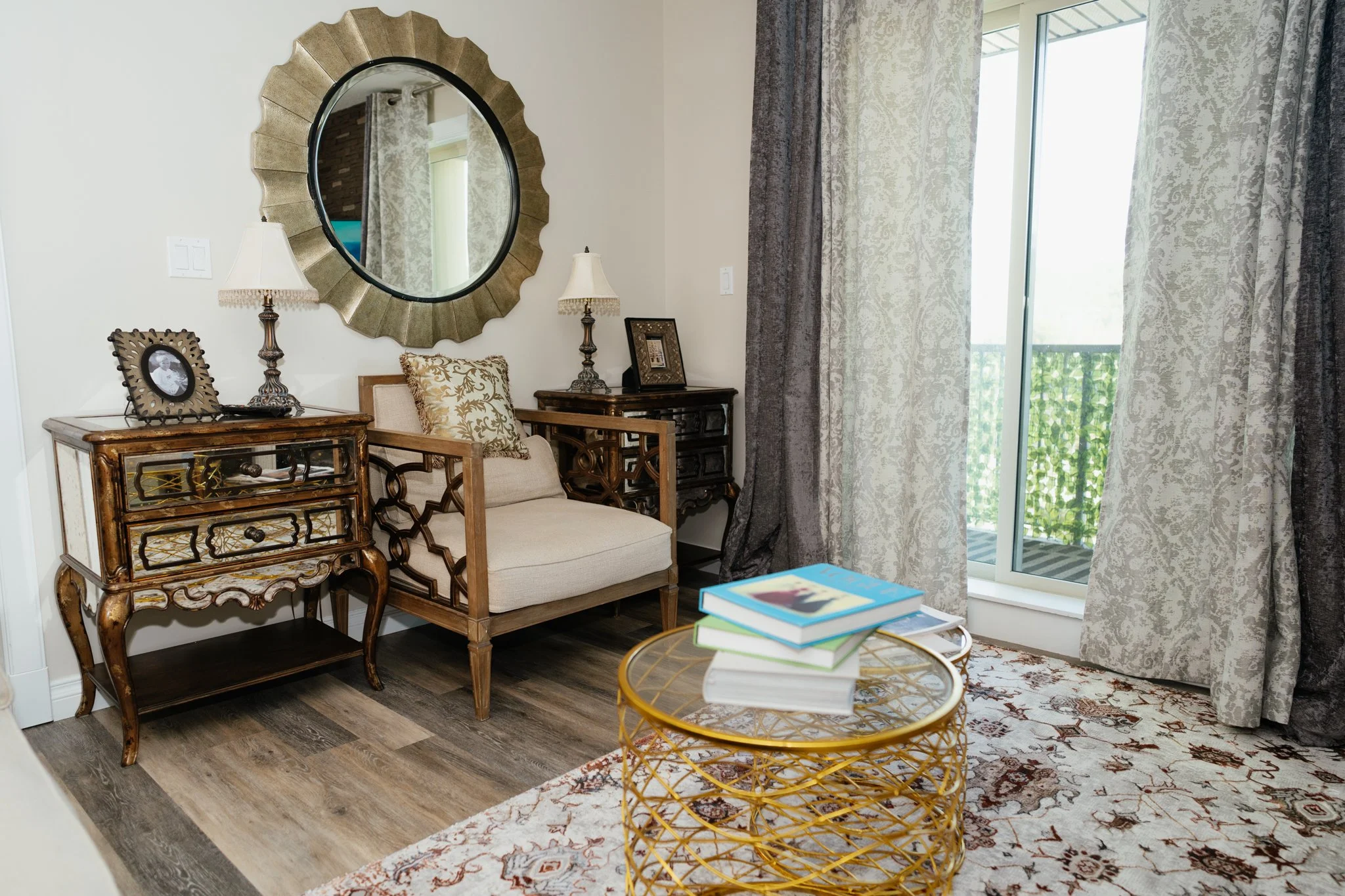 Living room with a large round mirror, lamps, a framed photo, armchair with patterned pillow, wooden side tables, a wire gold-colored coffee table, and sliding glass door with patterned curtains leading to a balcony.