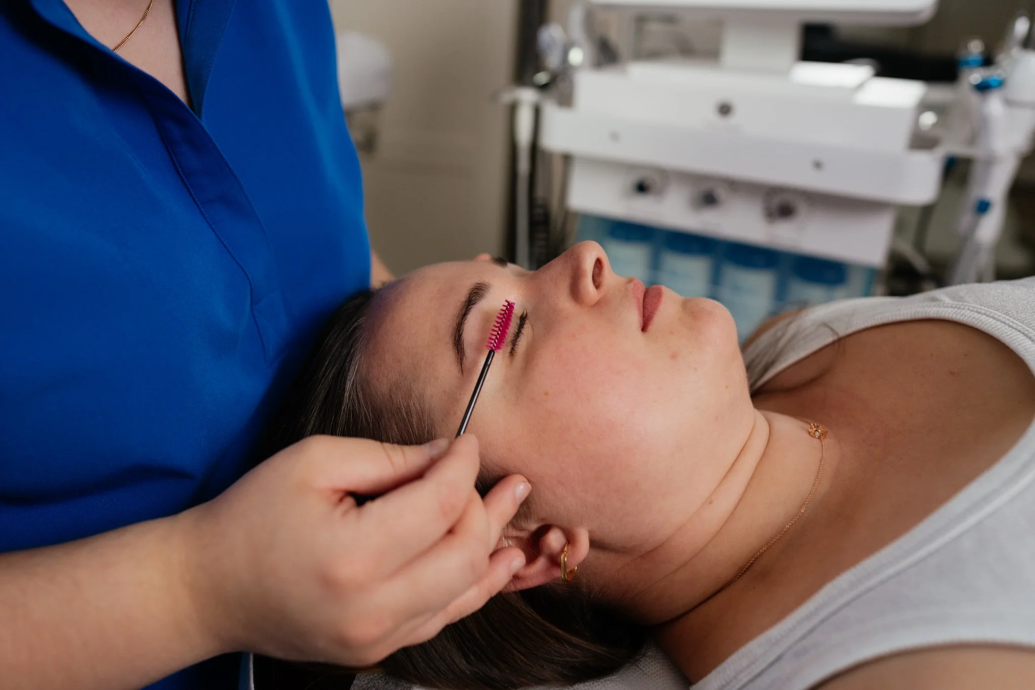 A woman lying on a treatment bed with her eyes closed, receiving eyelash extension or eyelash care from a professional. The professional is wearing a blue uniform and using a small tool near the woman's eye.
