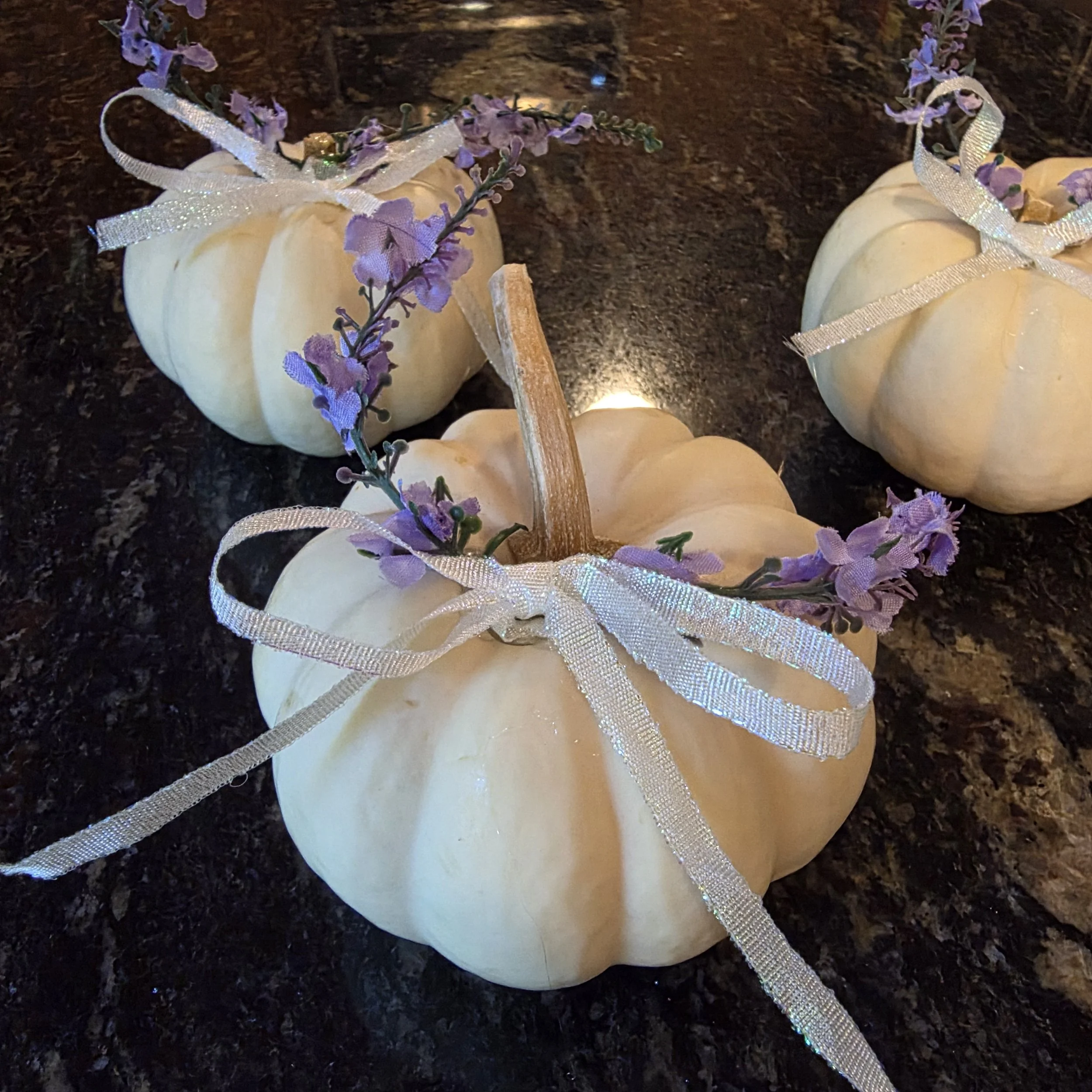 White pumpkins decorated with purple flowers, golden glitter, and silver ribbons are arranged on a dark surface.