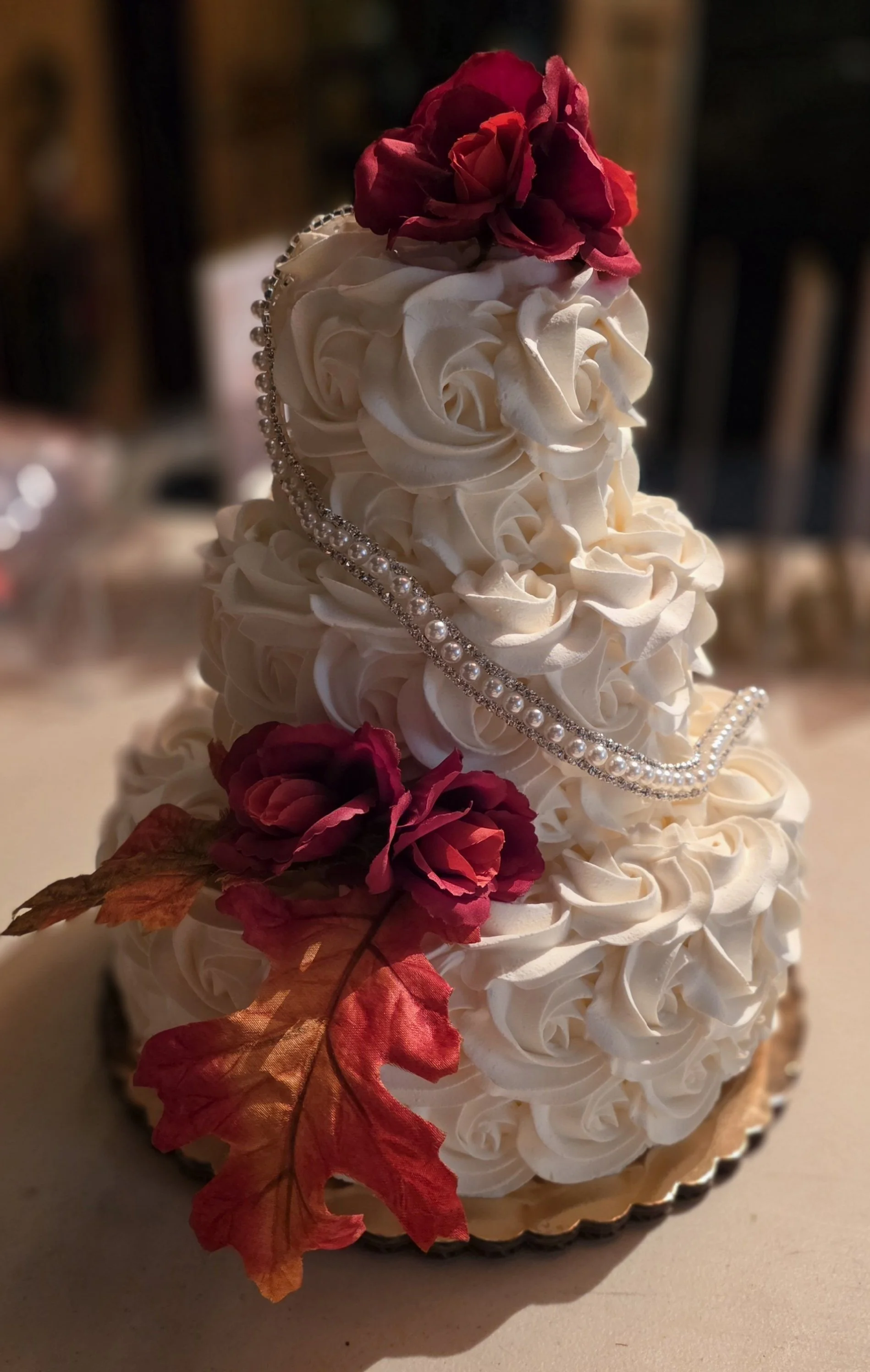 A dessert cake decorated with white frosting swirls, red and purple artificial flowers, a string of pearls, and a small cake at the base with autumn leaves.